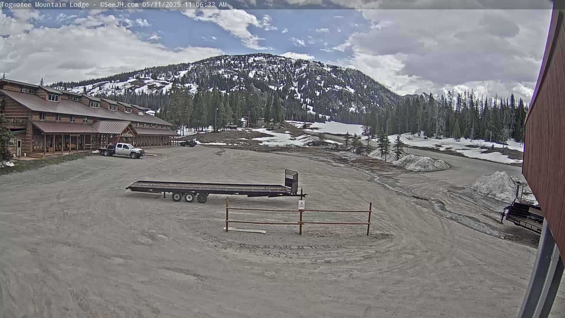 A large, rustic lodge sits in a parking lot with a flatbed trailer, partially snow-covered landscape, and mountains in the background under a partly cloudy sky.