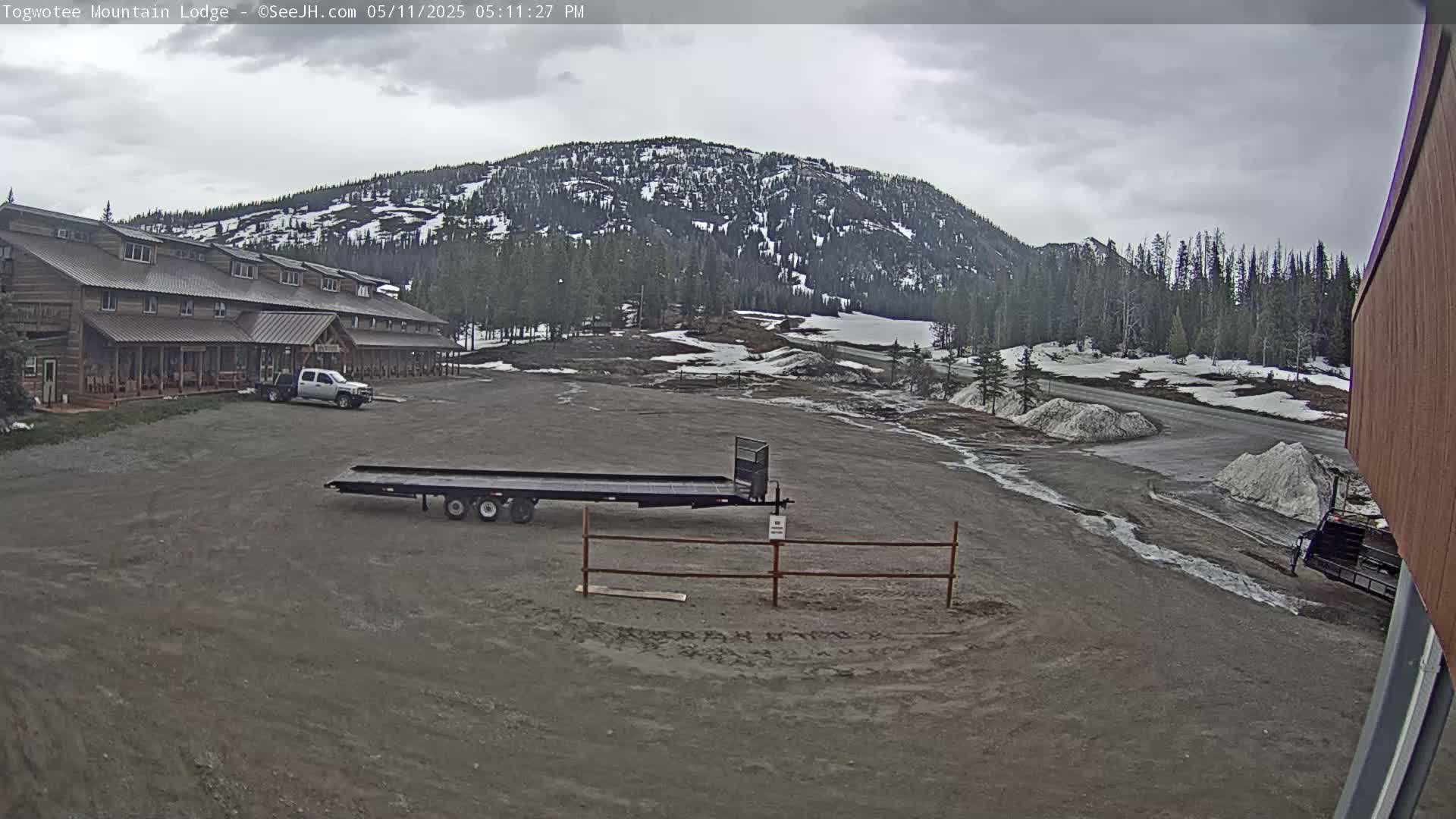 A large lodge sits in a parking lot with a flatbed trailer and truck, overlooking a mountain partially covered in snow under an overcast sky.
