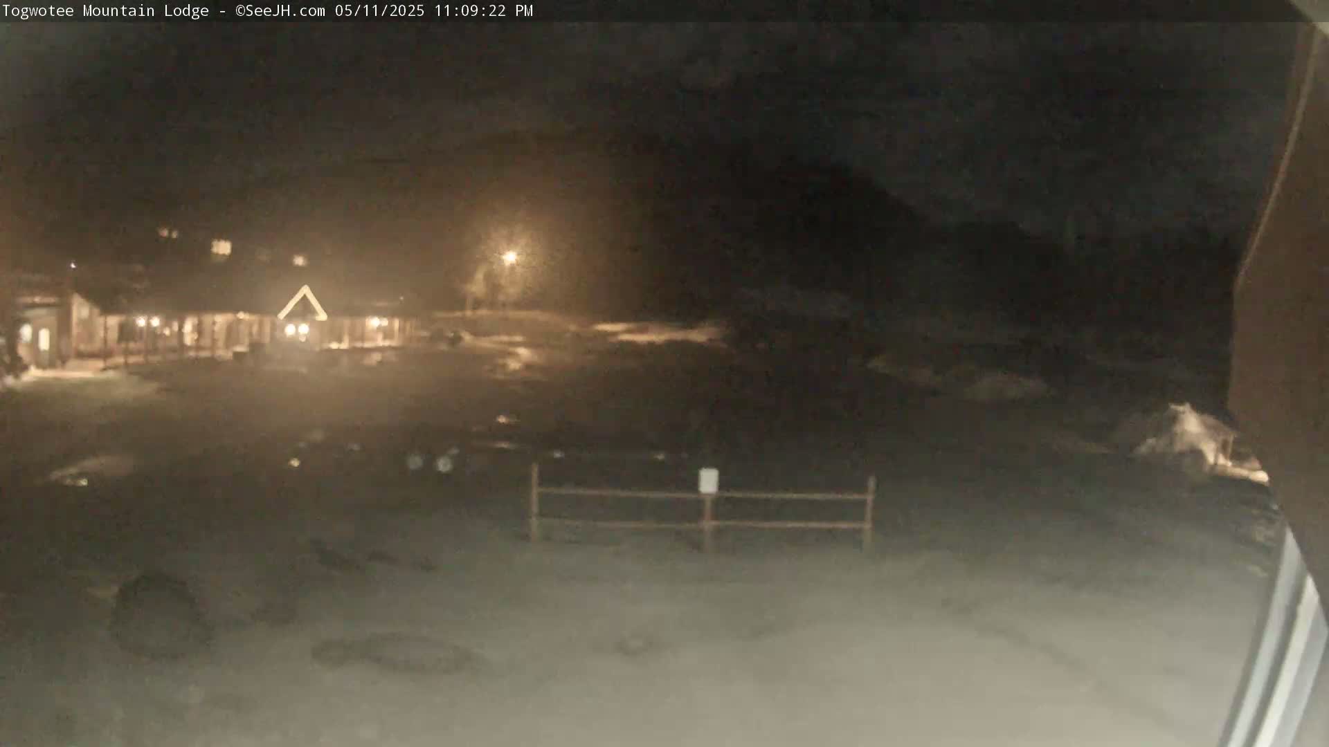 A nighttime view of a lodge, dimly lit parking lot, and a low fence, under a dark sky.
