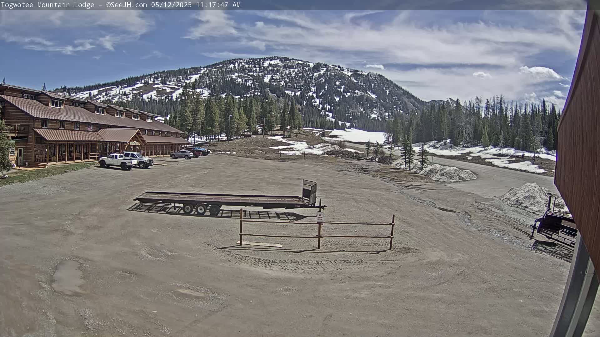 A large, log-style building with a parking lot in front sits below a snow-dusted mountain under a partly cloudy sky.