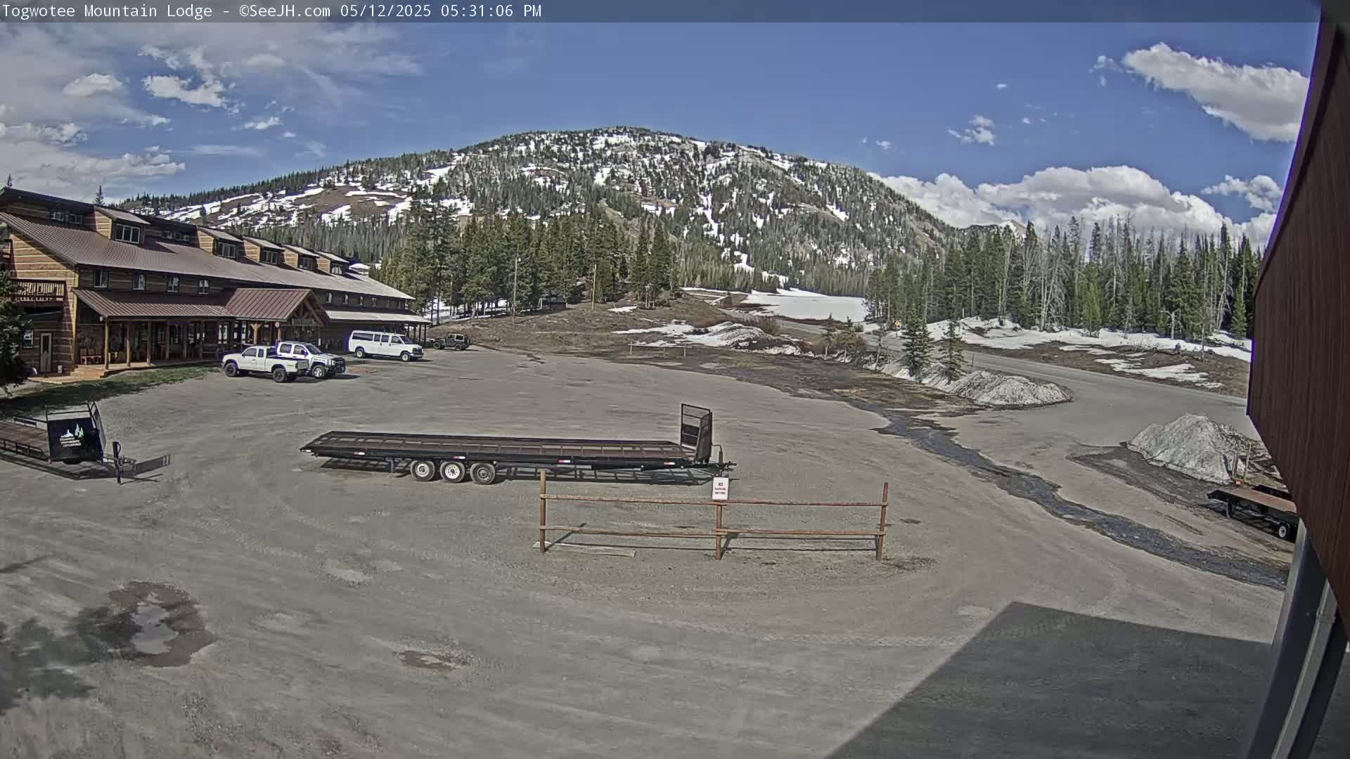 A large lodge sits near a mostly empty parking lot with several vehicles, trailers, and piles of what appears to be snow or gravel in front of a snow-capped mountain under a partly cloudy sky.