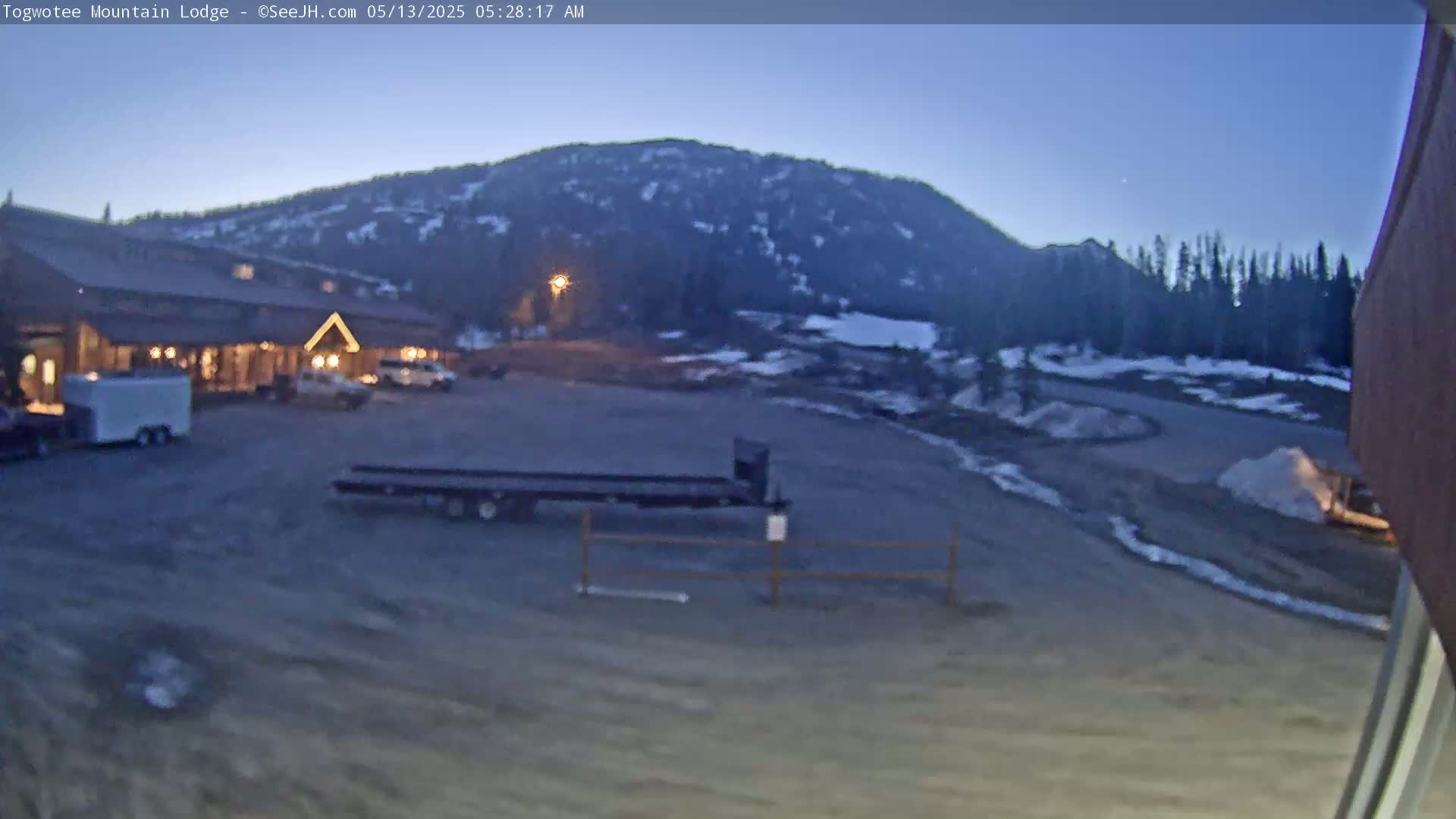 A lodge is situated in a parking lot under a twilight sky, with a mountain in the background and some remaining snow on the ground.