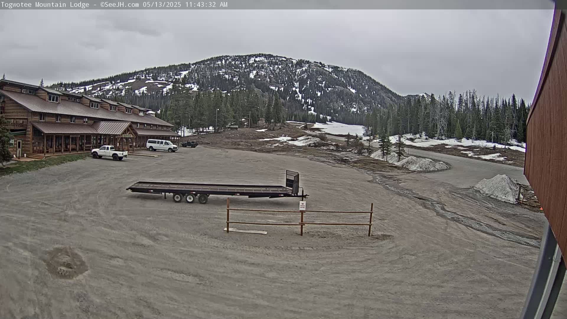 A large, brown lodge sits in a gravel parking lot in front of a snow-dusted mountain under an overcast sky.