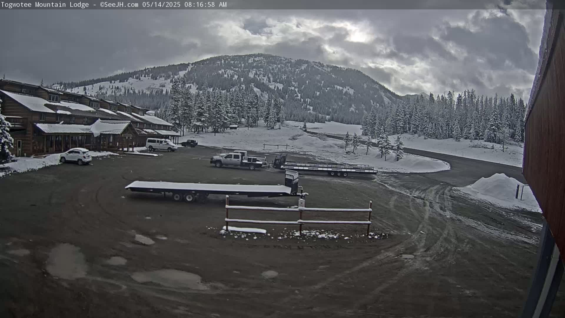 A snow-covered mountain and pine forest are visible behind a lodge and parking lot containing several vehicles and trailers; the sky is cloudy.