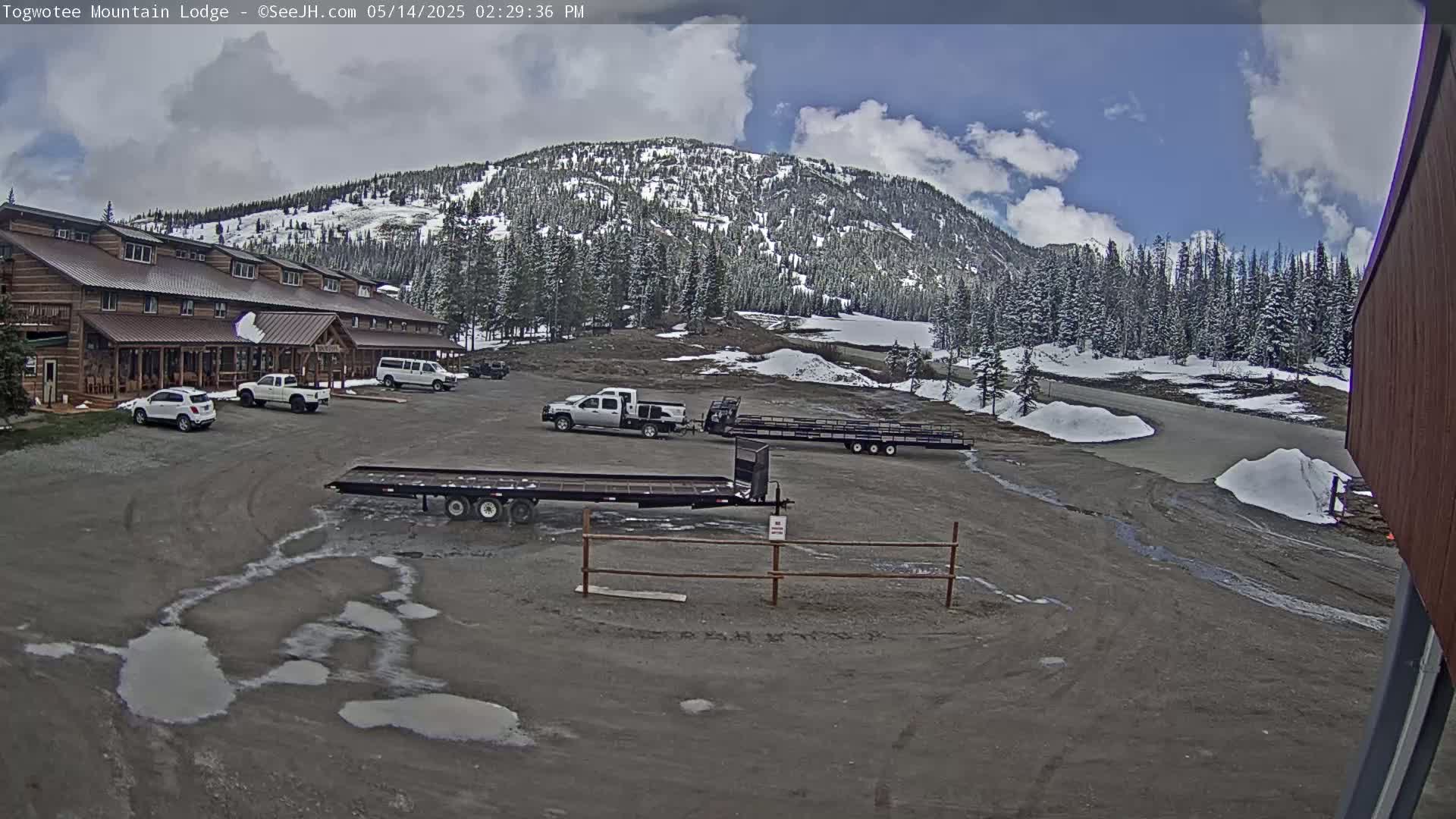 A lodge with several vehicles parked in front,  two flatbed trailers, and a partially snow-covered mountain in the background under a partly cloudy sky.