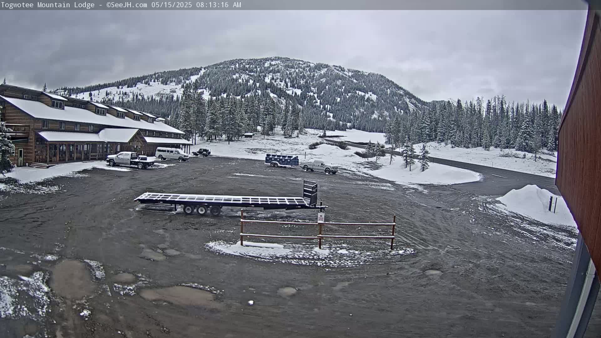 A snow-covered lodge and parking lot are situated in front of a snow-dusted mountain range under an overcast sky.