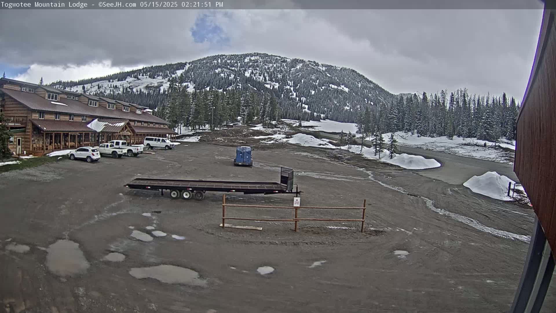 A lodge with several cars parked in front sits in a muddy parking lot overlooking a snow-dusted mountain under a cloudy sky.