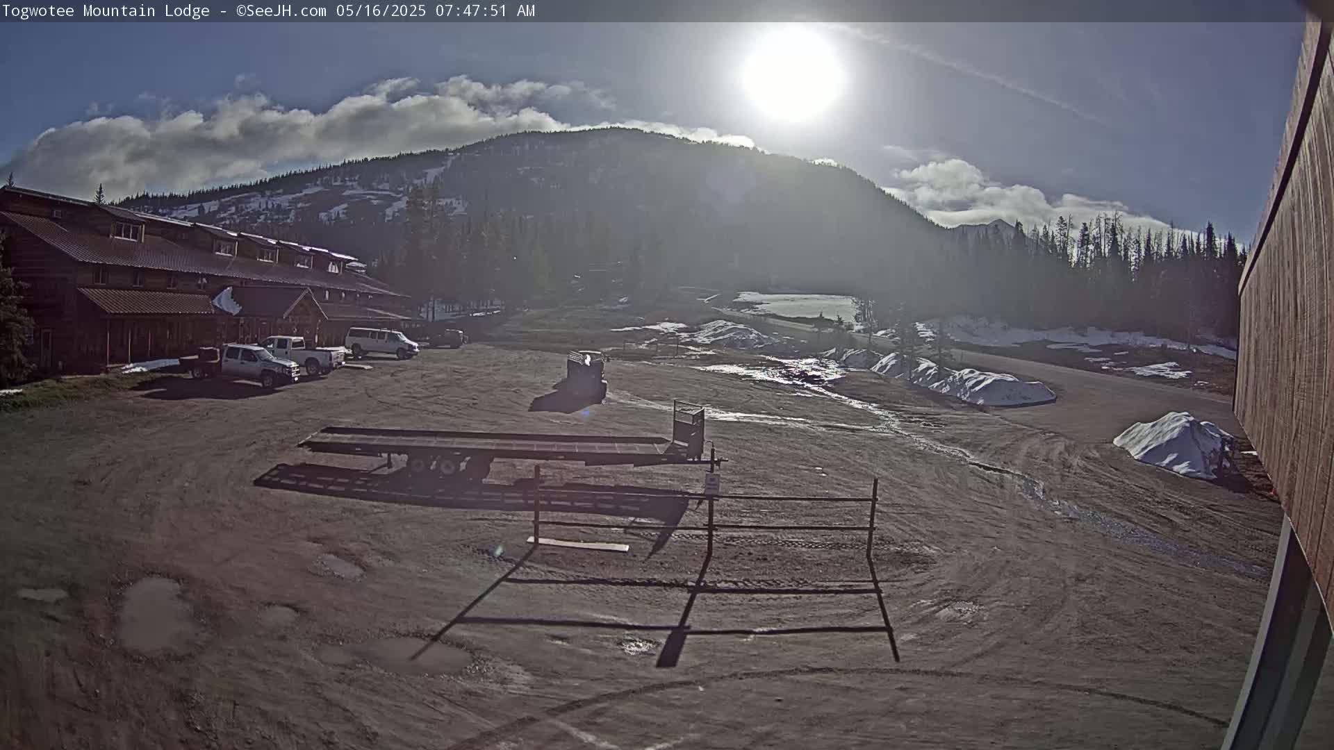 A sunny day reveals a lodge with a parking lot containing several vehicles, a flatbed trailer, and small piles of snow, all situated before a mountain under a partly cloudy sky.