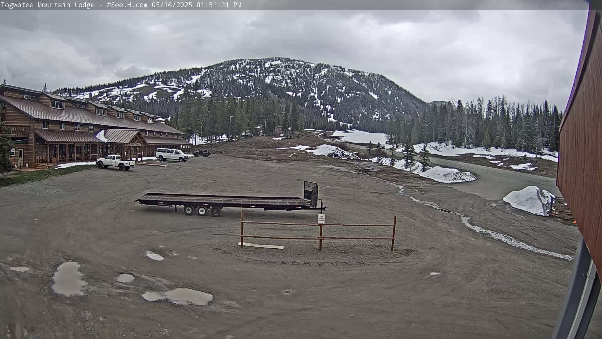 A large, rustic lodge sits in a gravel parking lot under an overcast sky, with a snow-dusted mountain in the background.