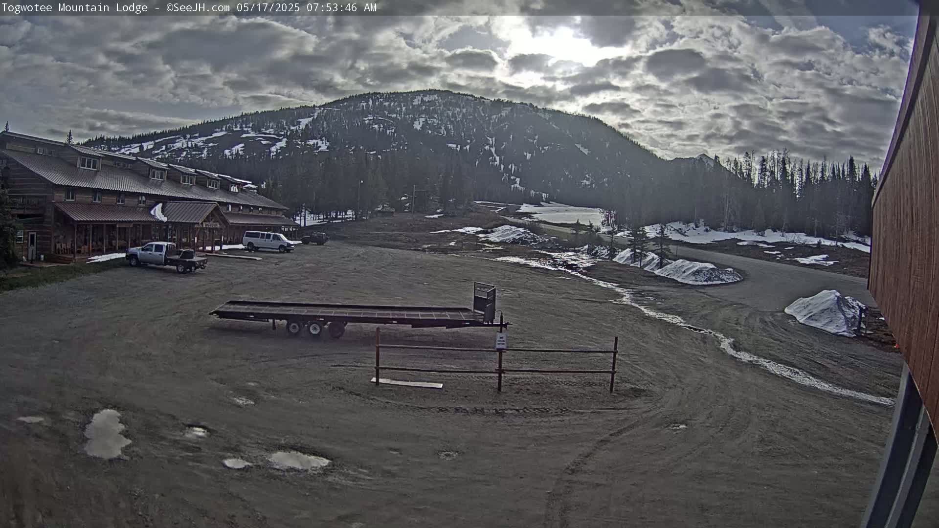 A large lodge sits in a parking lot, with a few vehicles visible, under a partly cloudy sky; a mountain is visible in the background, with some patches of snow.