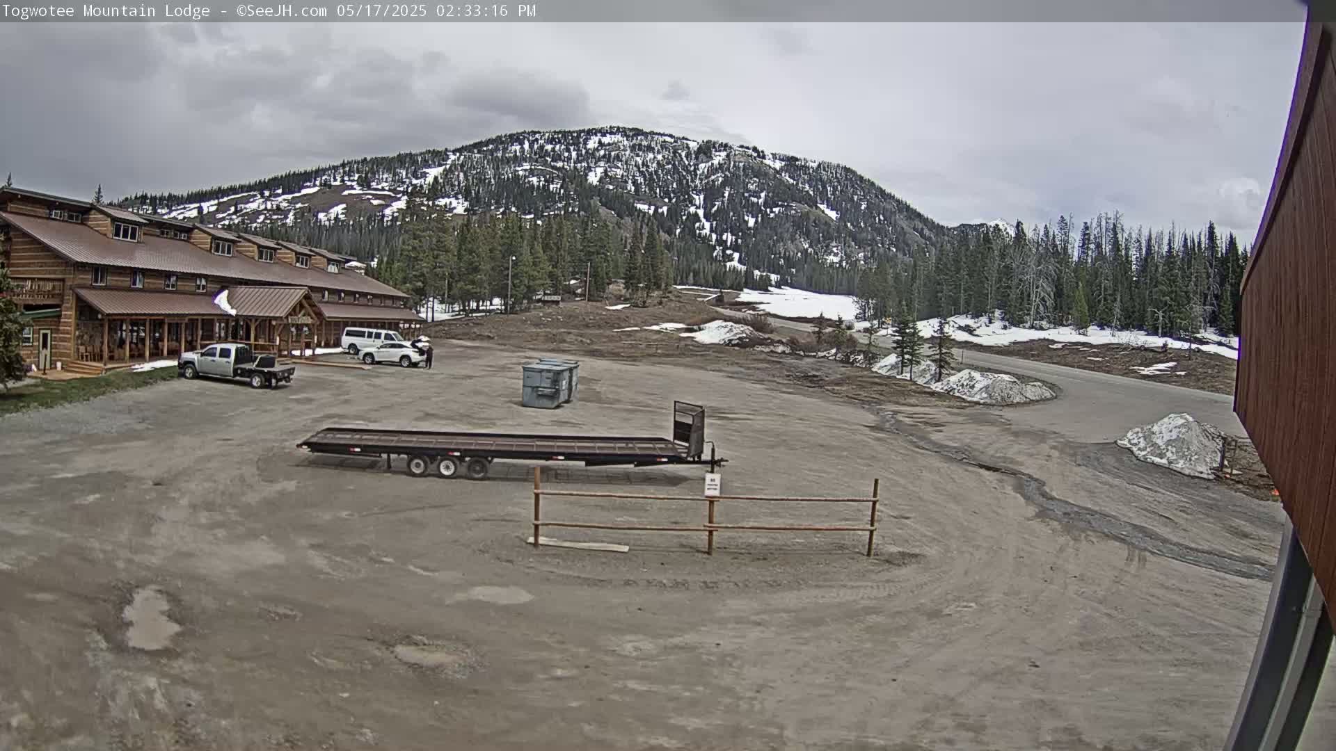 A large, rustic lodge is situated in a parking lot with a few vehicles, a flatbed trailer, and dumpsters, all under an overcast sky, with snow-capped mountains and a coniferous forest in the background.