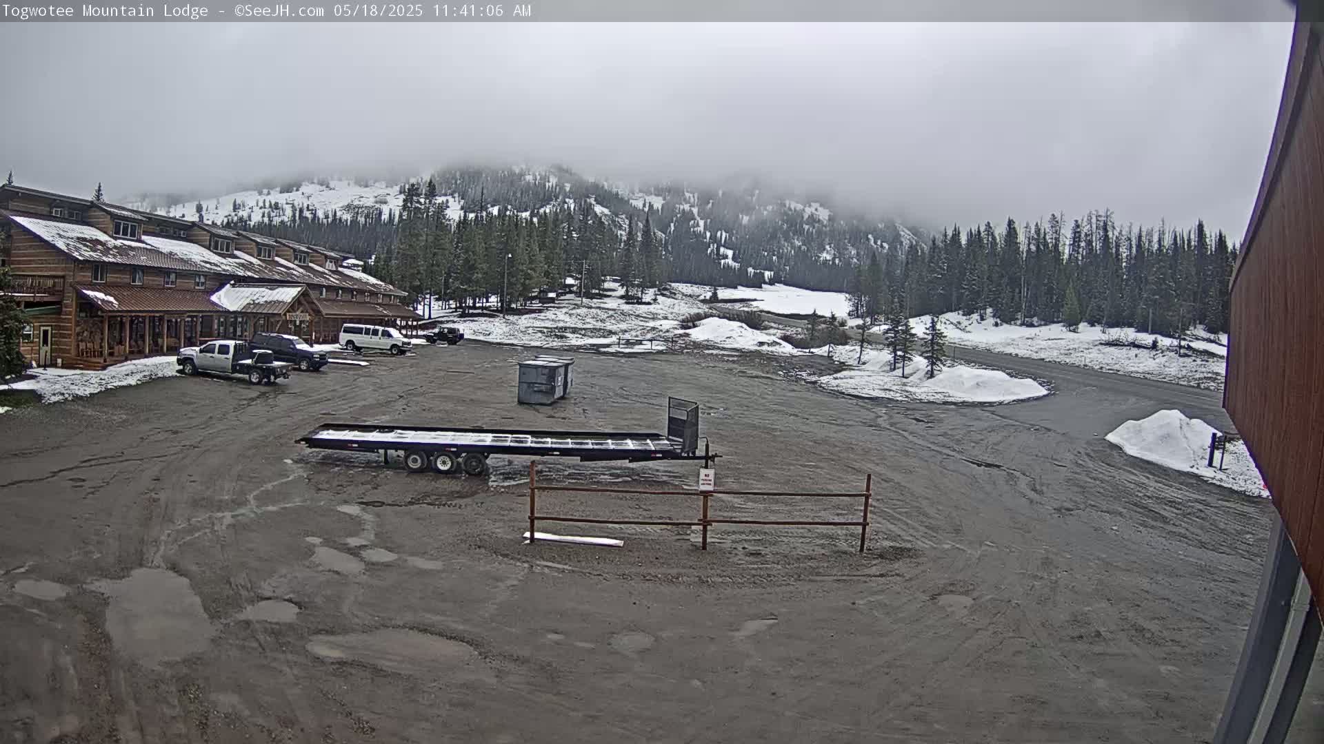 A mostly cloudy day reveals a snow-dusted parking lot in front of a log cabin building, with a flatbed trailer and several vehicles parked nearby, and a mountainous background partially obscured by fog.