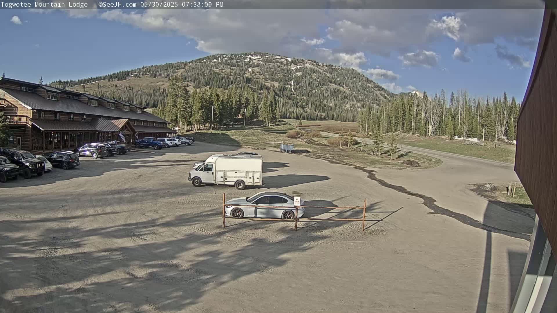 A large, multi-story log building with a parking lot full of cars and a van is situated in front of a mountain under a partly cloudy, sunny sky.
