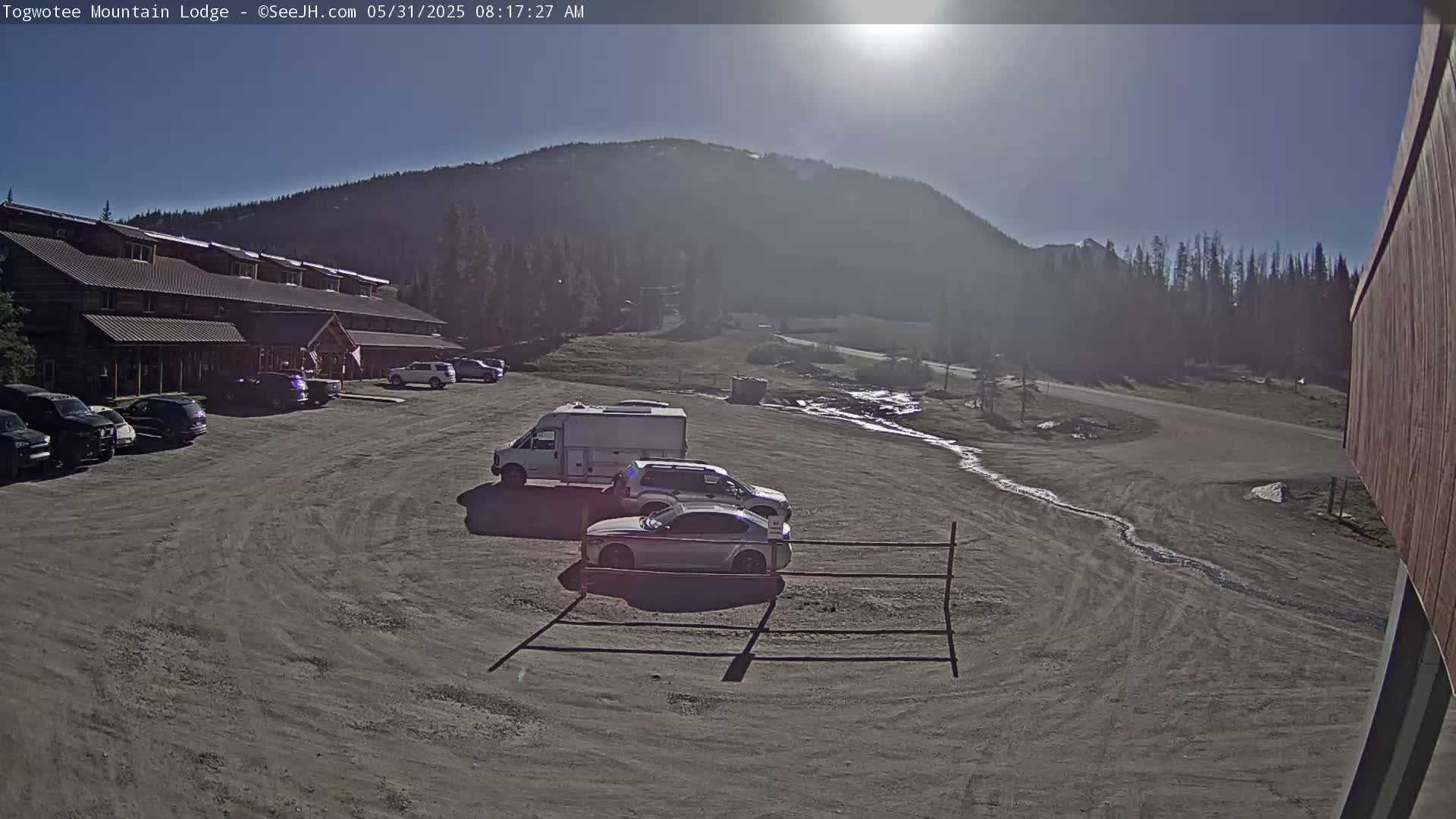 A sunny day reveals a parking lot with several cars and a van in front of a long, low building nestled against a mountain backdrop.