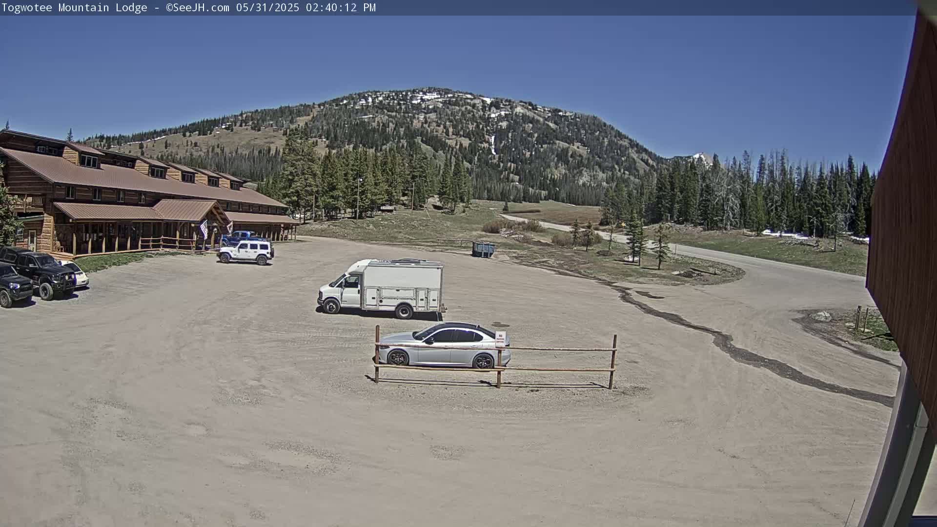 A large, brown lodge sits in a parking lot with several vehicles, under a clear blue sky in front of a mountain covered in trees and some patches of snow.
