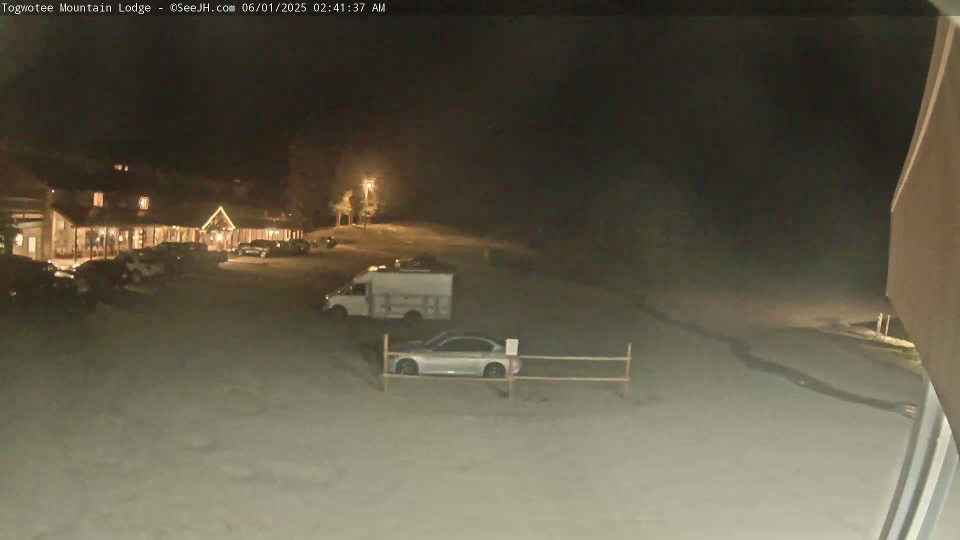 A nighttime view of a lodge parking lot with several cars and a van parked, under dark, clear skies.