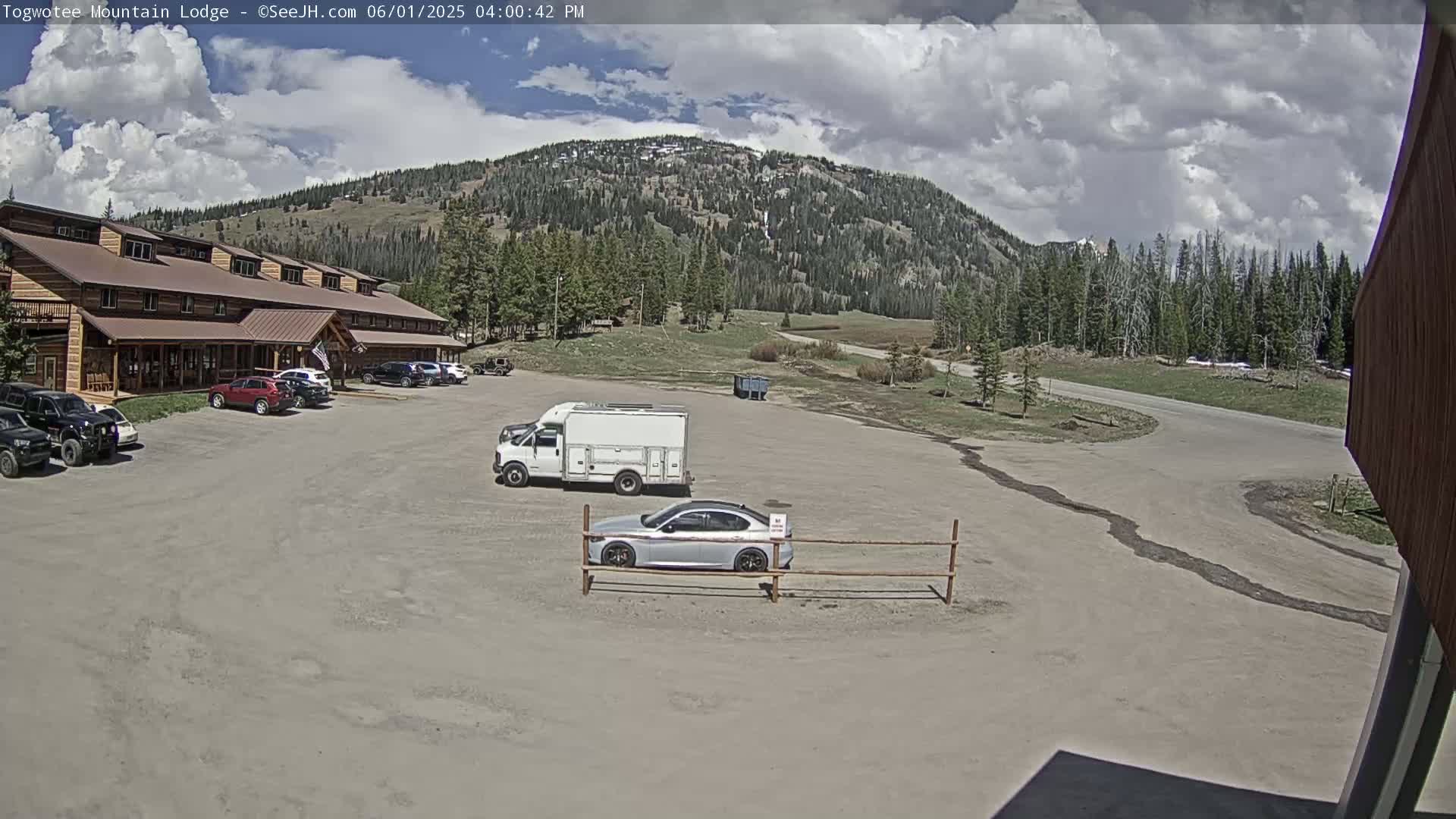 A large, log-style building with several cars parked in front, a few other vehicles in a gravel parking lot, and a mountain with partly cloudy skies in the background.
