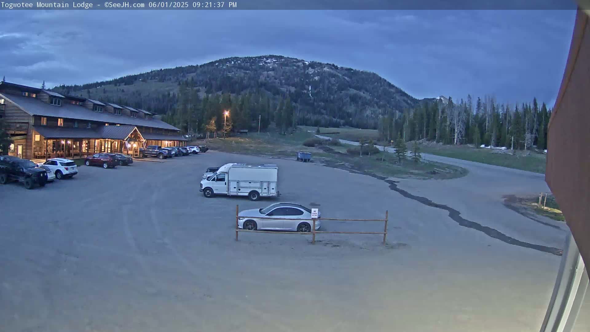 A large lodge sits in a parking lot with several cars and a van, under a partly cloudy twilight sky, with a mountain and trees in the background.