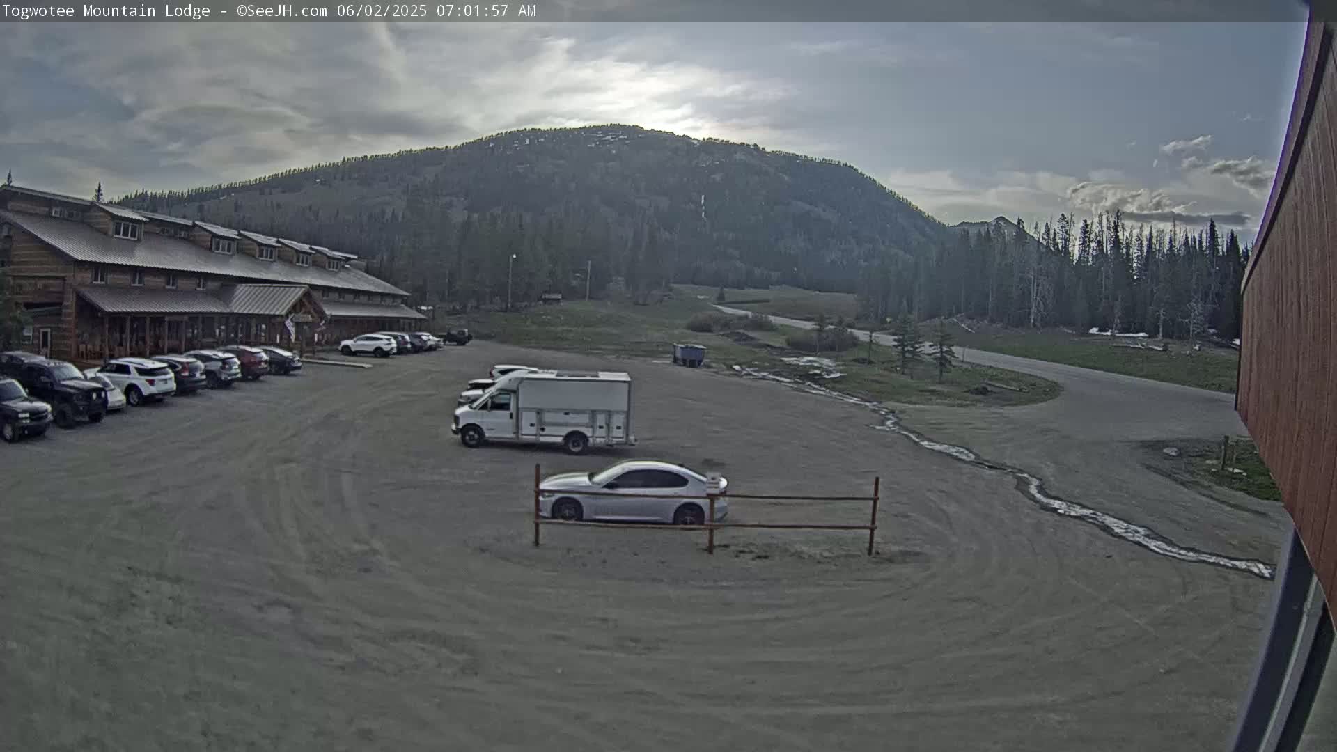 A parking lot with several cars and a van in front of a long, log cabin-style building, with a mountain and forest in the background under a mostly cloudy sky.
