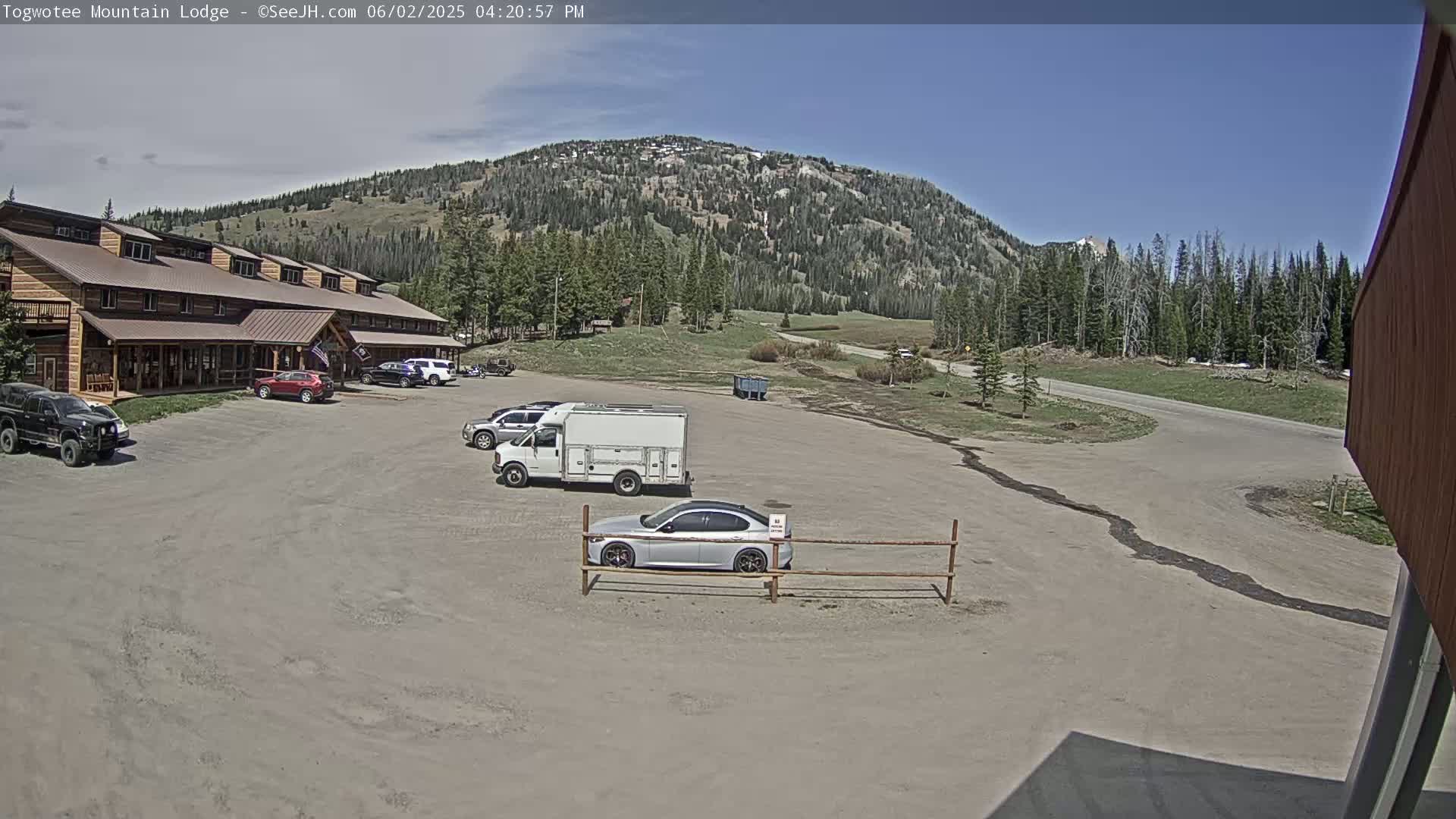 A lodge with a parking lot containing several vehicles is situated in front of a mountain range under a mostly clear sky.