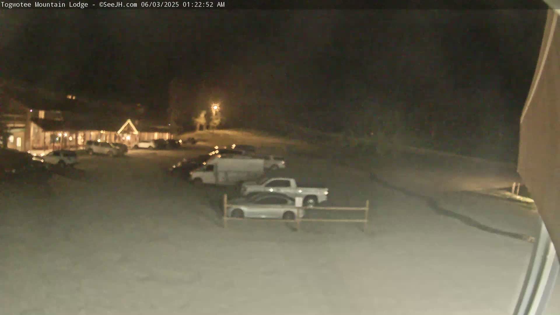 A nighttime, snow-covered parking lot with several vehicles parked near a lodge.