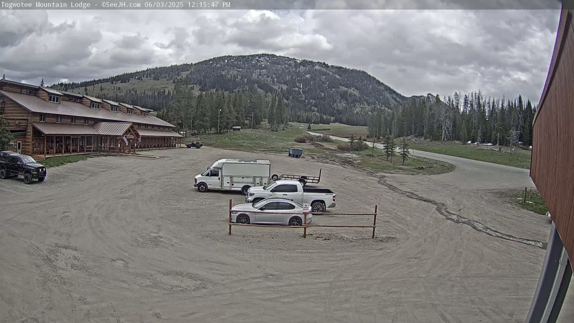 A large, long, log cabin-style building sits in a gravel parking lot with several vehicles, under a cloudy sky, with a mountain in the background.