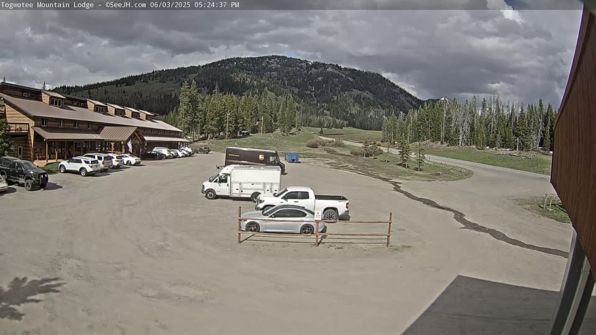 A parking lot with several cars and trucks in front of a lodge, under a partly cloudy sky, with a mountain in the background.