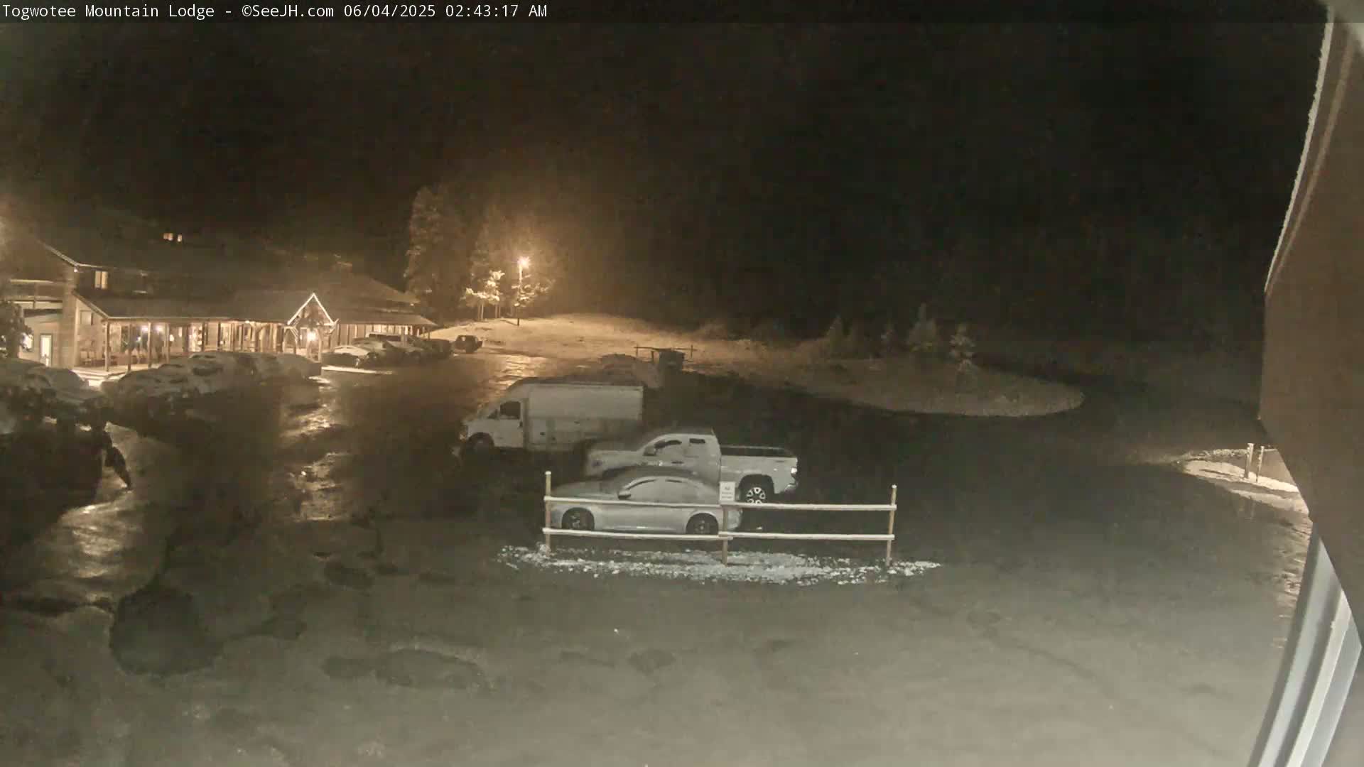 A nighttime, snowy scene shows a lodge with several vehicles parked in a lot, some covered in snow.