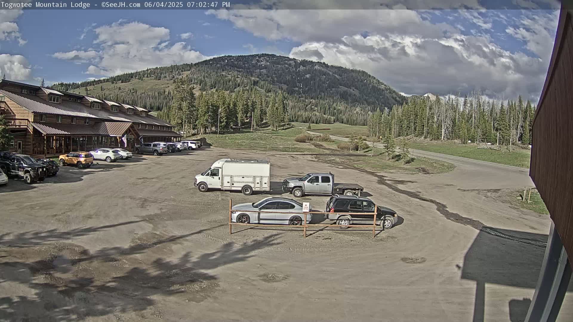 A lodge parking lot with several vehicles, including a van and pickup truck, sits in front of a large, rustic-style building with a mountain backdrop under partly cloudy skies.