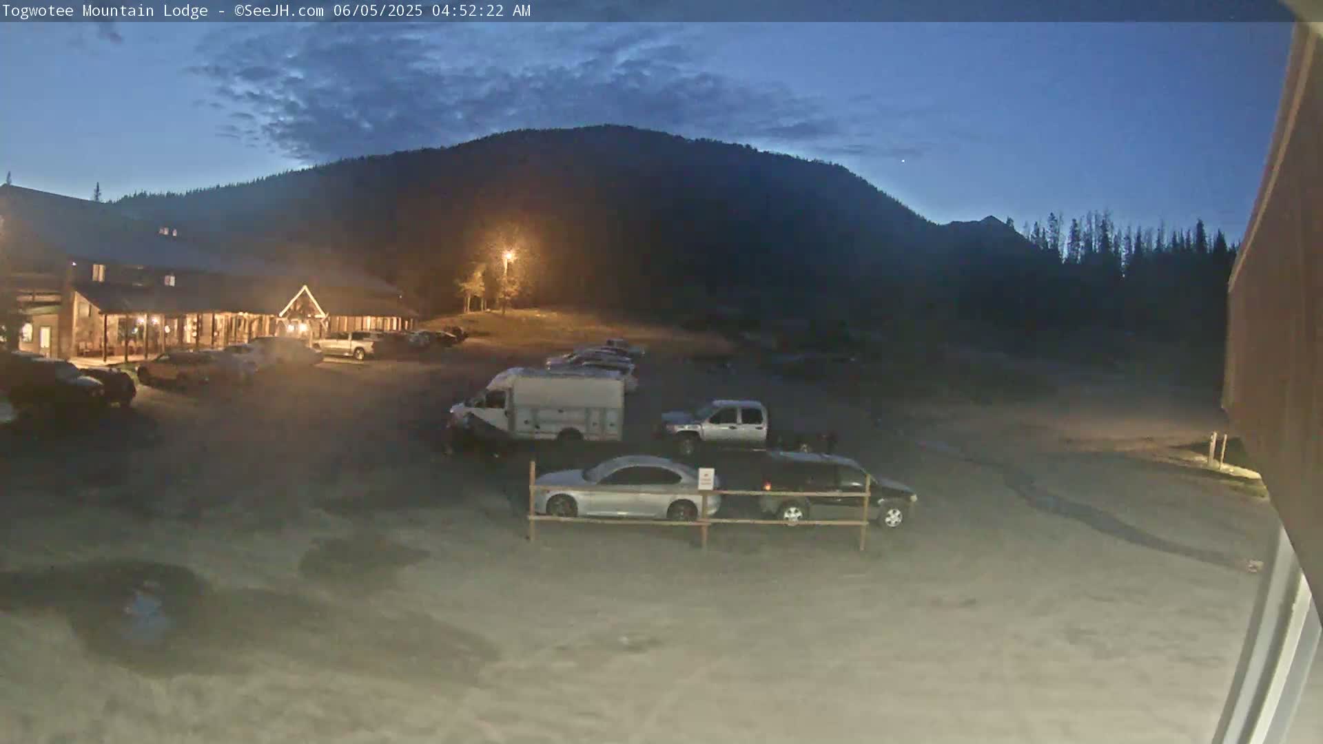 A lodge is illuminated at twilight under a mostly clear sky, with a parking lot containing several vehicles in front of it and a mountain behind it.
