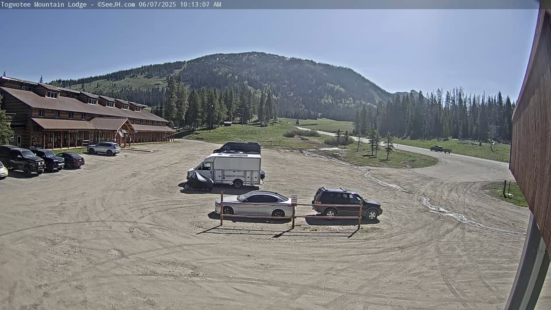 A lodge with several cars parked in front, a van and an SUV in the parking lot, and a mountain in the background under a clear, sunny sky.