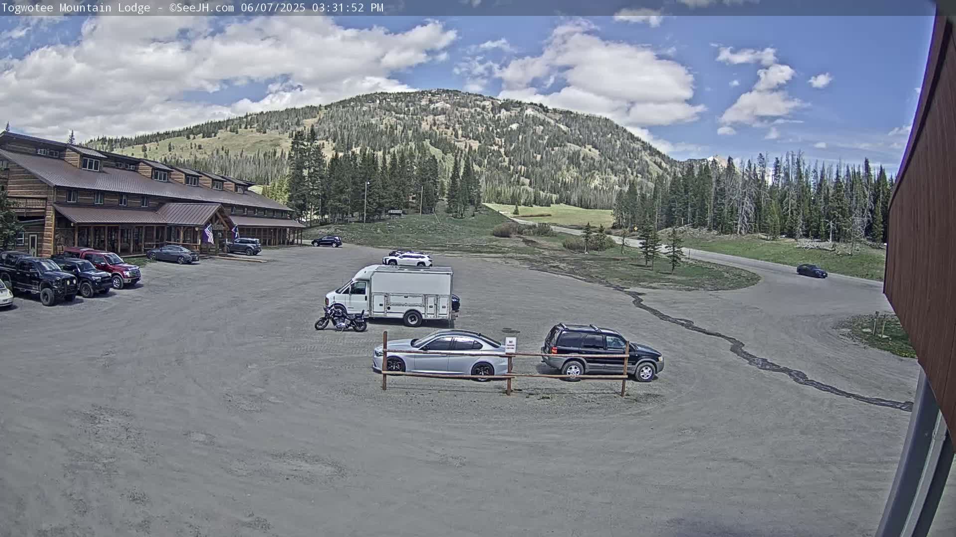 A large lodge sits in a parking lot with several cars and a van, overlooking a mountain range under a partly cloudy sky.