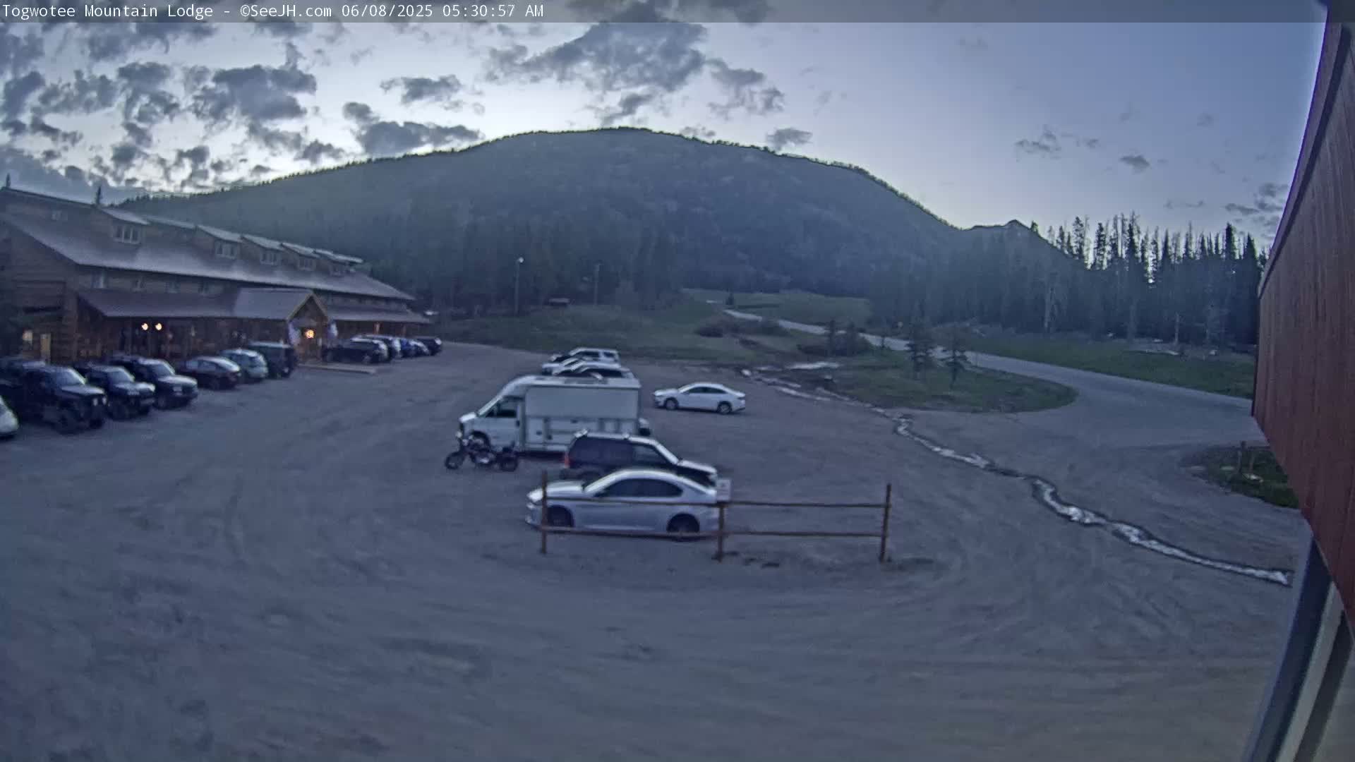 A lodge with a parking lot filled with cars and a camper van sits in front of a mountain under an overcast sky.