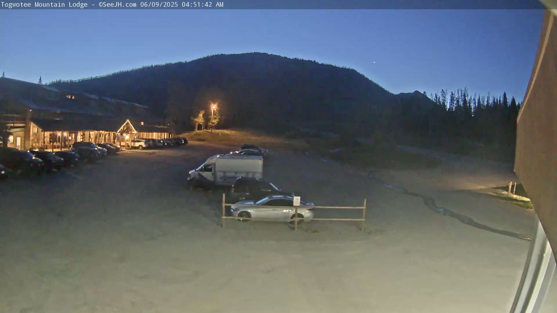 A lit lodge with a parking lot full of cars sits before a dark mountain under a twilight sky.