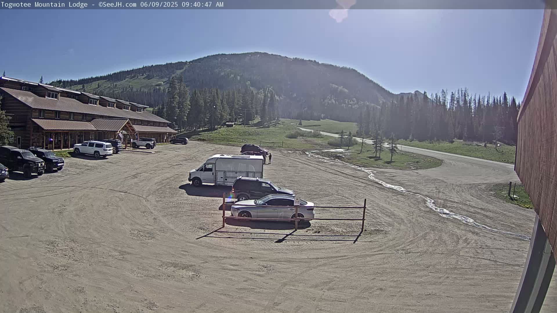 A large, wood-sided lodge is situated in a parking lot with several vehicles, overlooking a grassy valley and mountain under a clear, sunny sky.