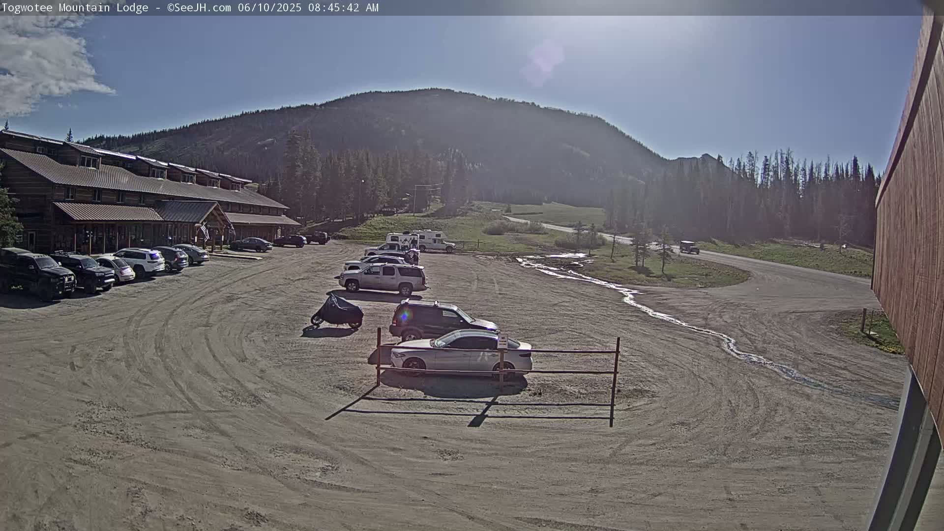 A lodge with a parking lot full of cars sits below a mountain on a sunny day.