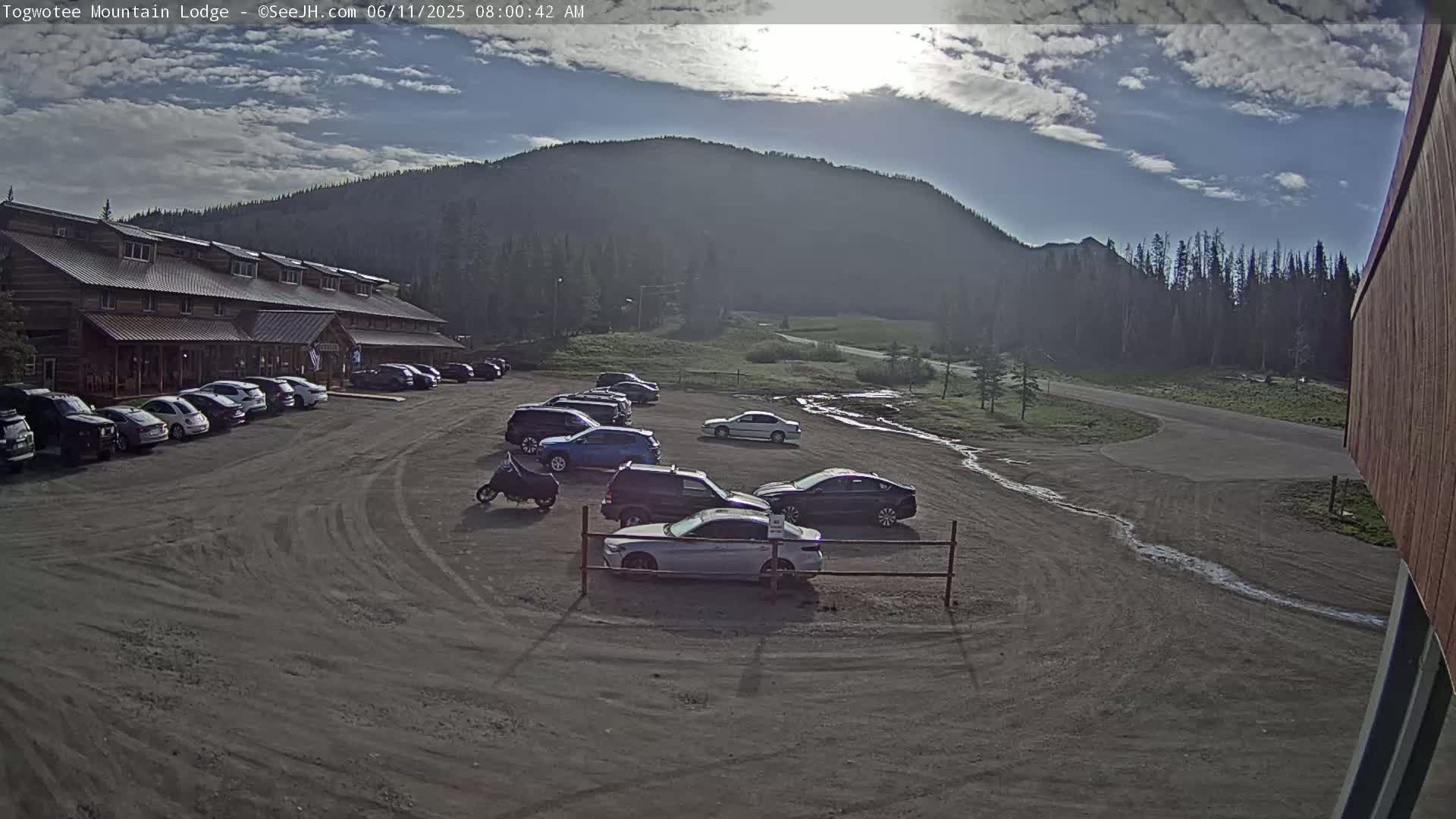 A parking lot with several cars parked in front of a long, log cabin-style building, under a mostly sunny sky with some clouds and a mountain in the background.