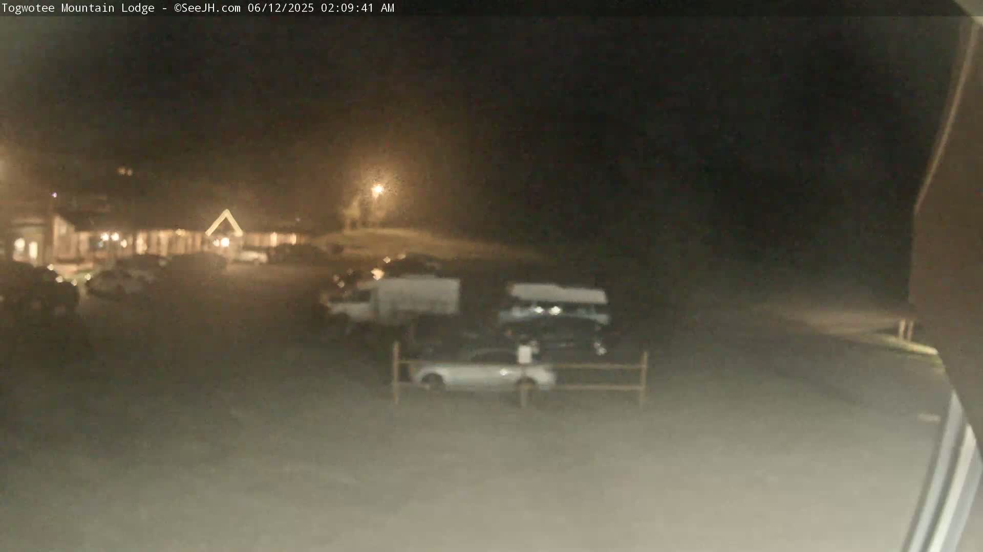 A nighttime, blurry view of a parking lot with several cars and vans in front of a building under dim lighting.