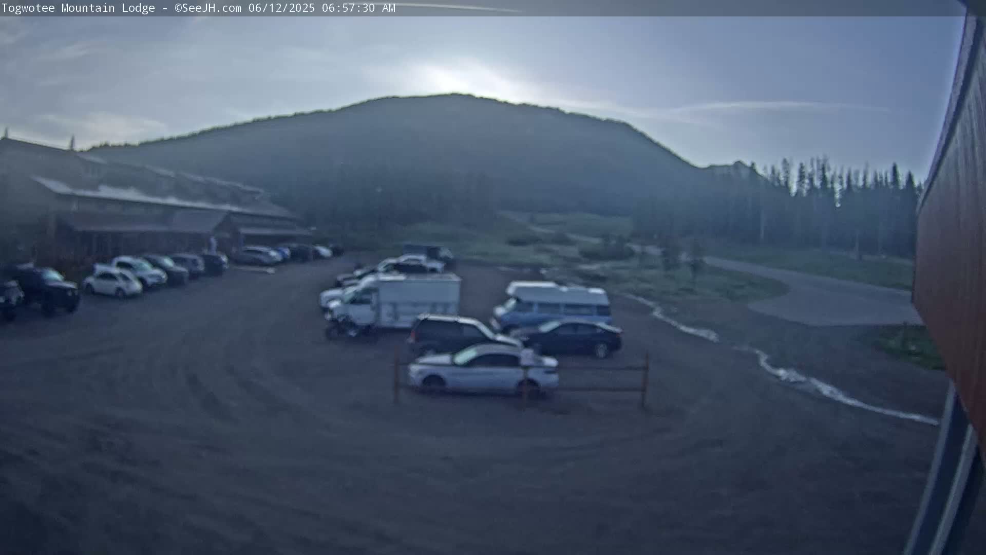 A parking lot with several cars and RVs is situated in front of a lodge, with a hazy mountain visible in the background under a mostly clear sky.