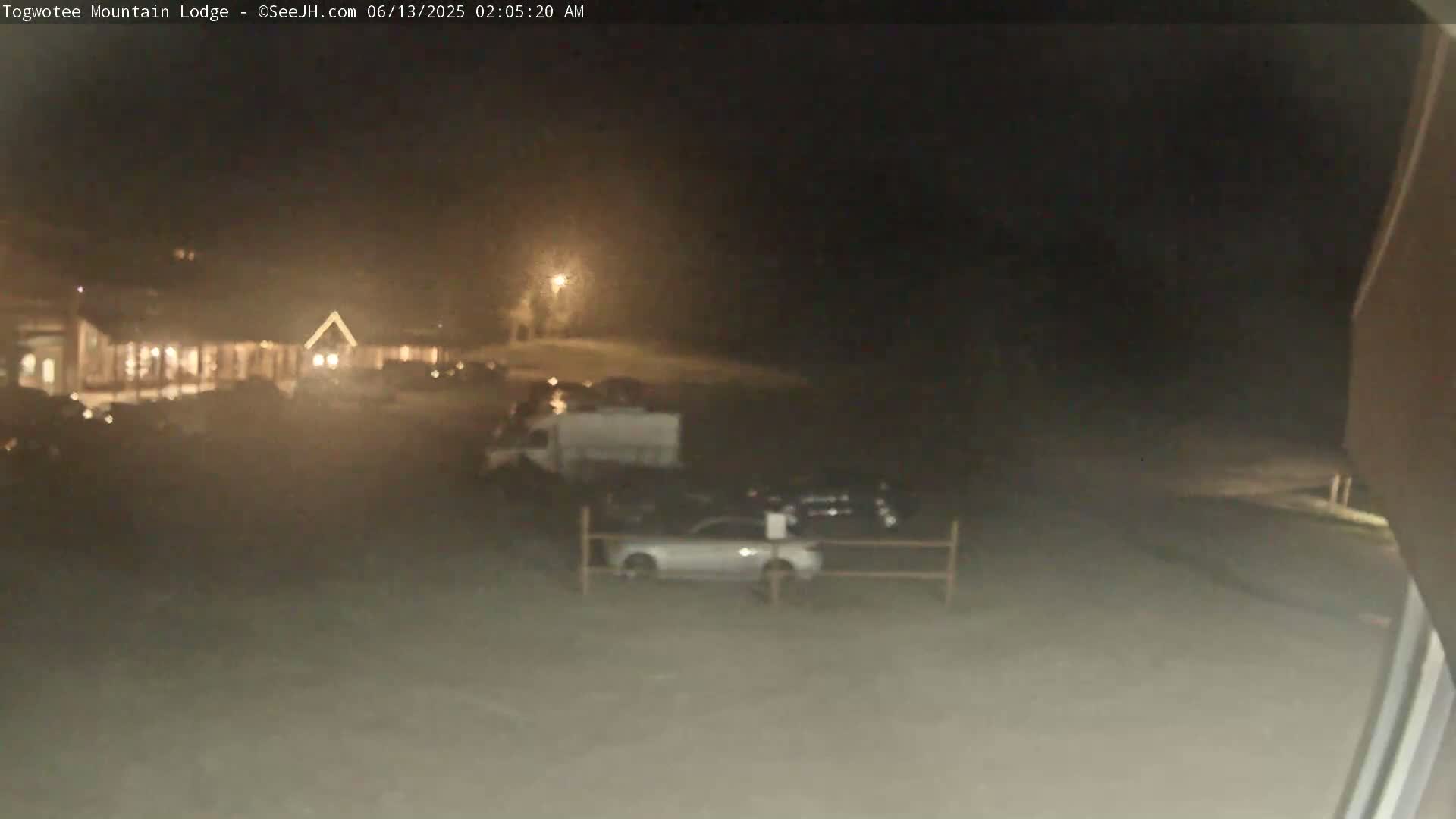 A nighttime, hazy view shows a parking lot with several cars and a van parked in front of a building with lights on.