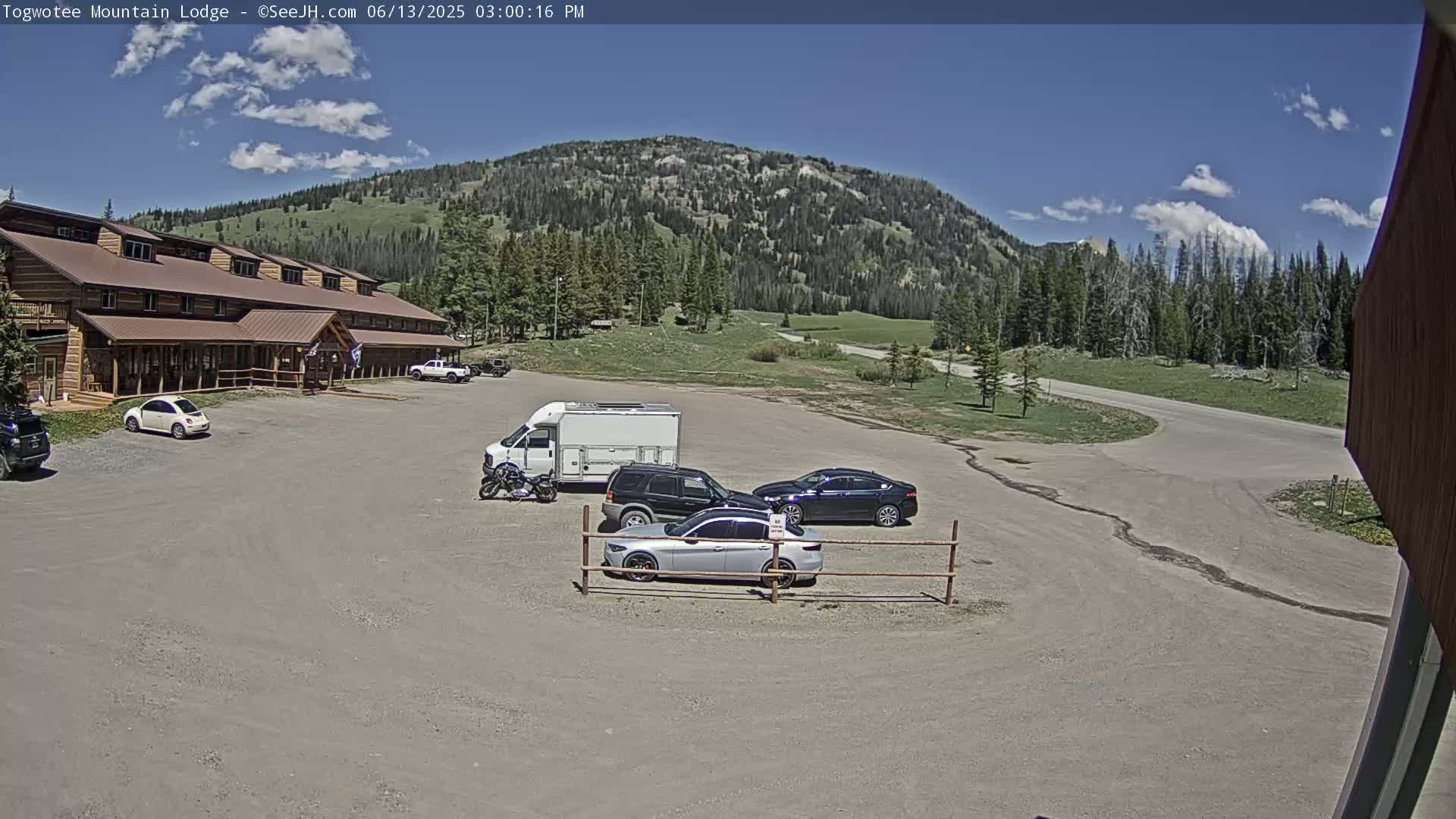 A long, brown lodge sits in a gravel parking lot with several cars and a camper van parked nearby, under a mostly sunny sky with some scattered clouds, in front of a large green mountain.