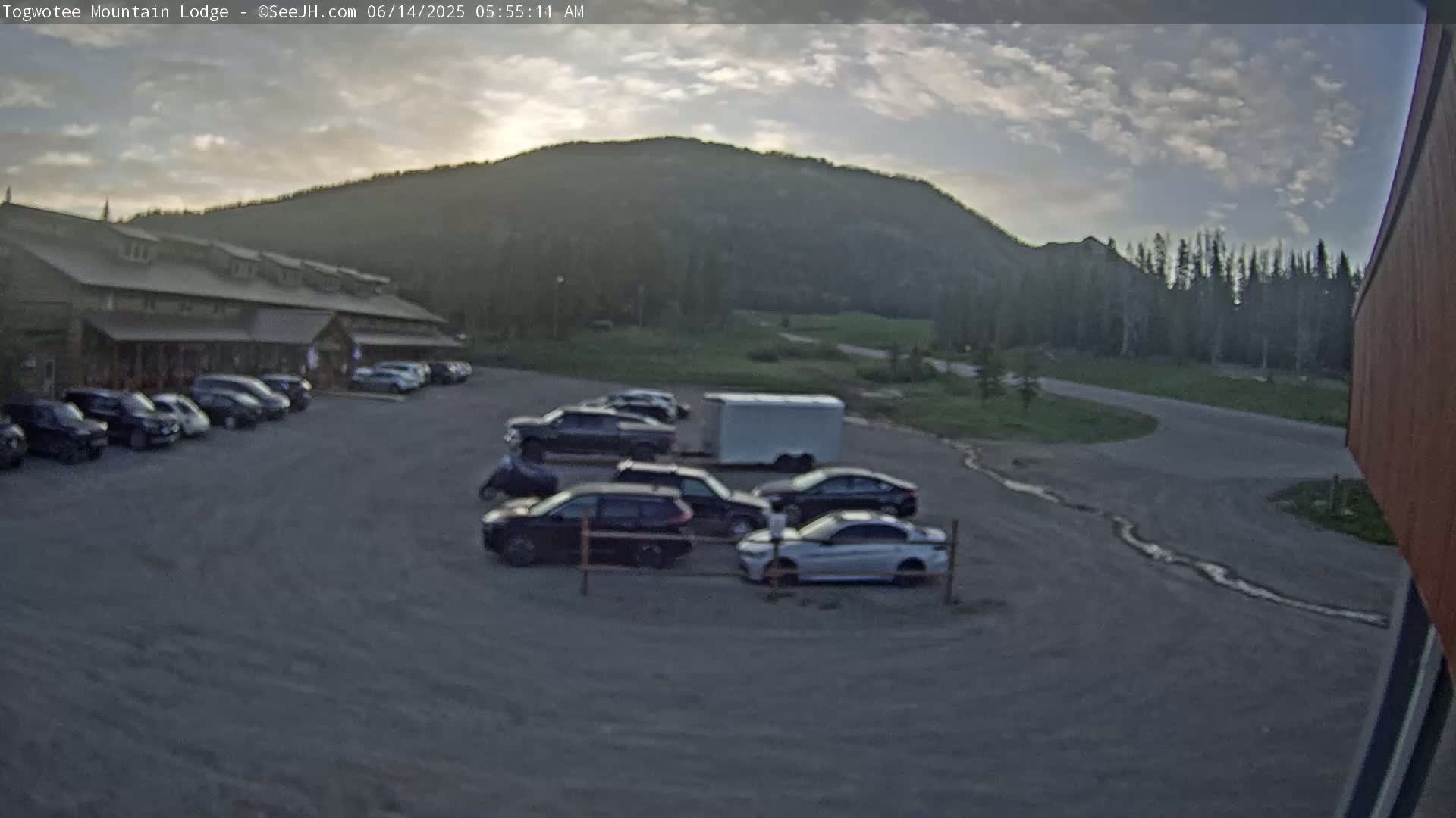 A parking lot with several cars parked in front of a long building, situated below a hill under a partly cloudy sky.