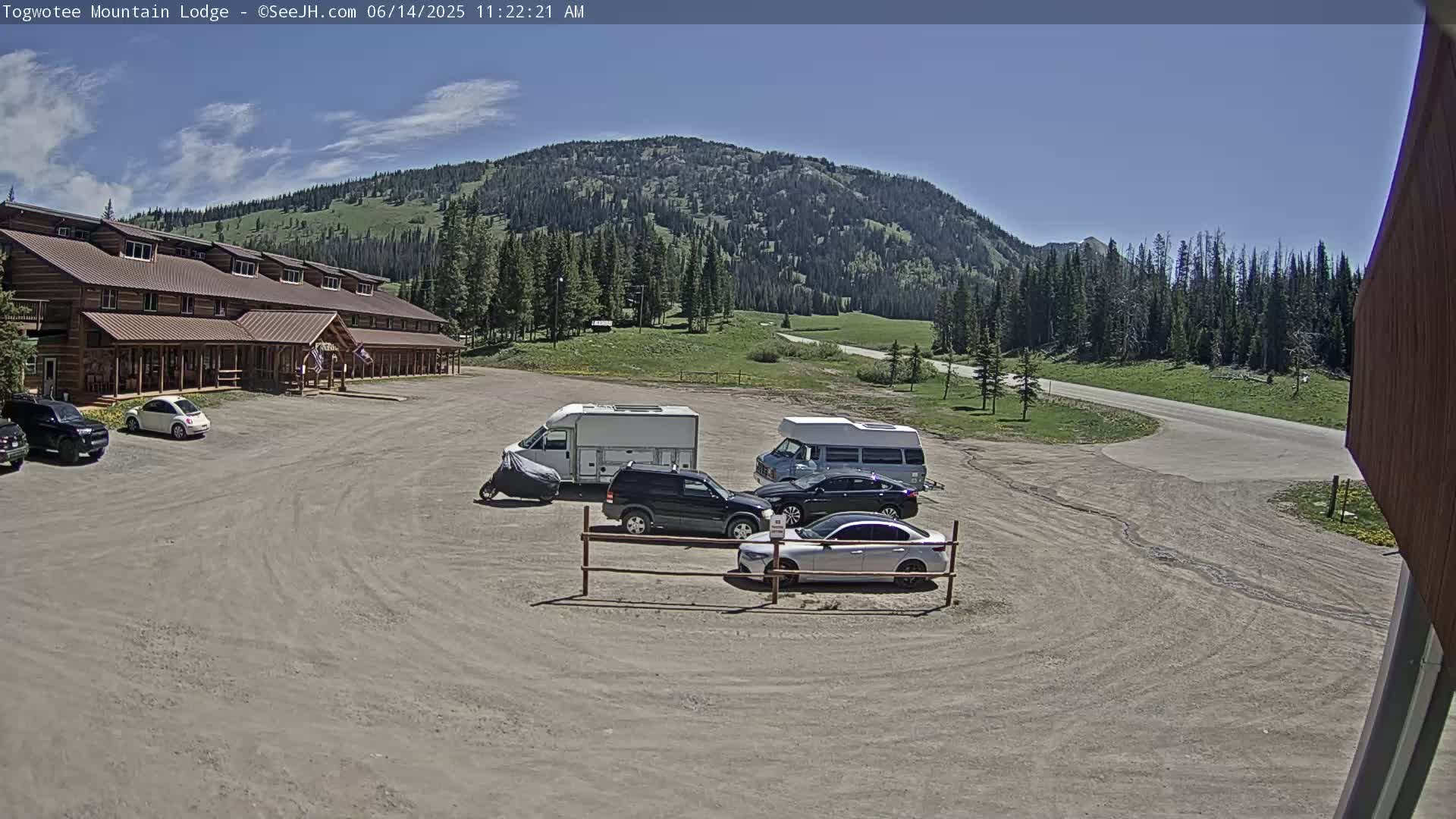 A long, brown lodge sits in a gravel parking lot with several vehicles, including cars and camper vans, under a clear blue sky in front of a mountain range.