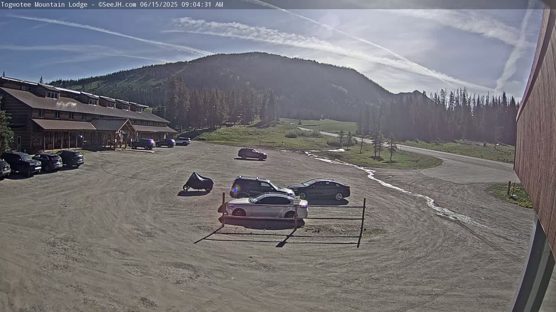 A lodge with several cars parked in front, under a mostly sunny sky with some clouds, is situated in a mountainous area with trees and a road visible in the background.