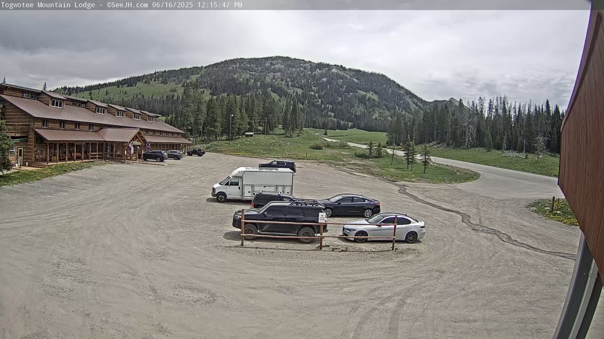 A lodge sits in a gravel parking lot with several vehicles parked nearby, all under a partly cloudy sky, with a mountain and green hills visible in the background.