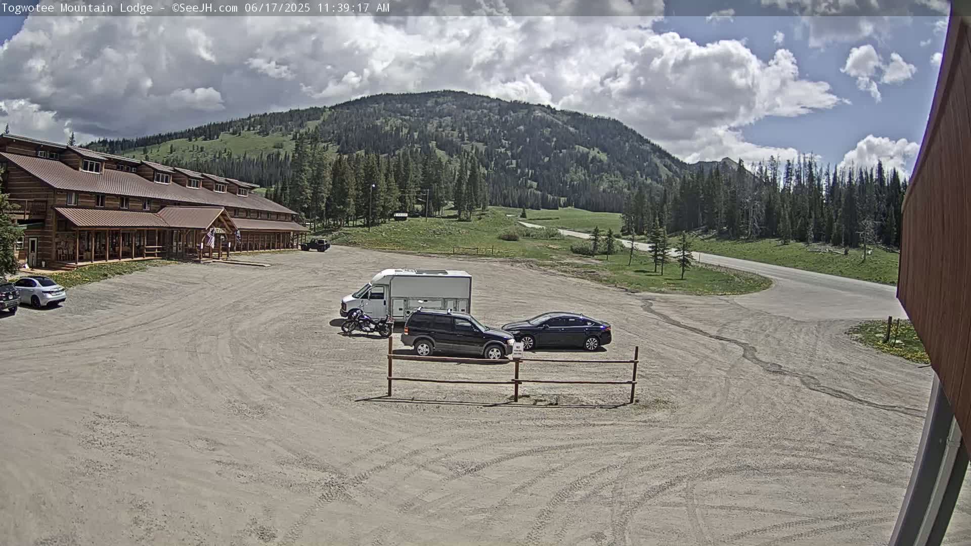 A long, log cabin-style building sits in a gravel parking lot with several vehicles, including an SUV and a campervan, under a partly cloudy sky and in front of a mountain range.