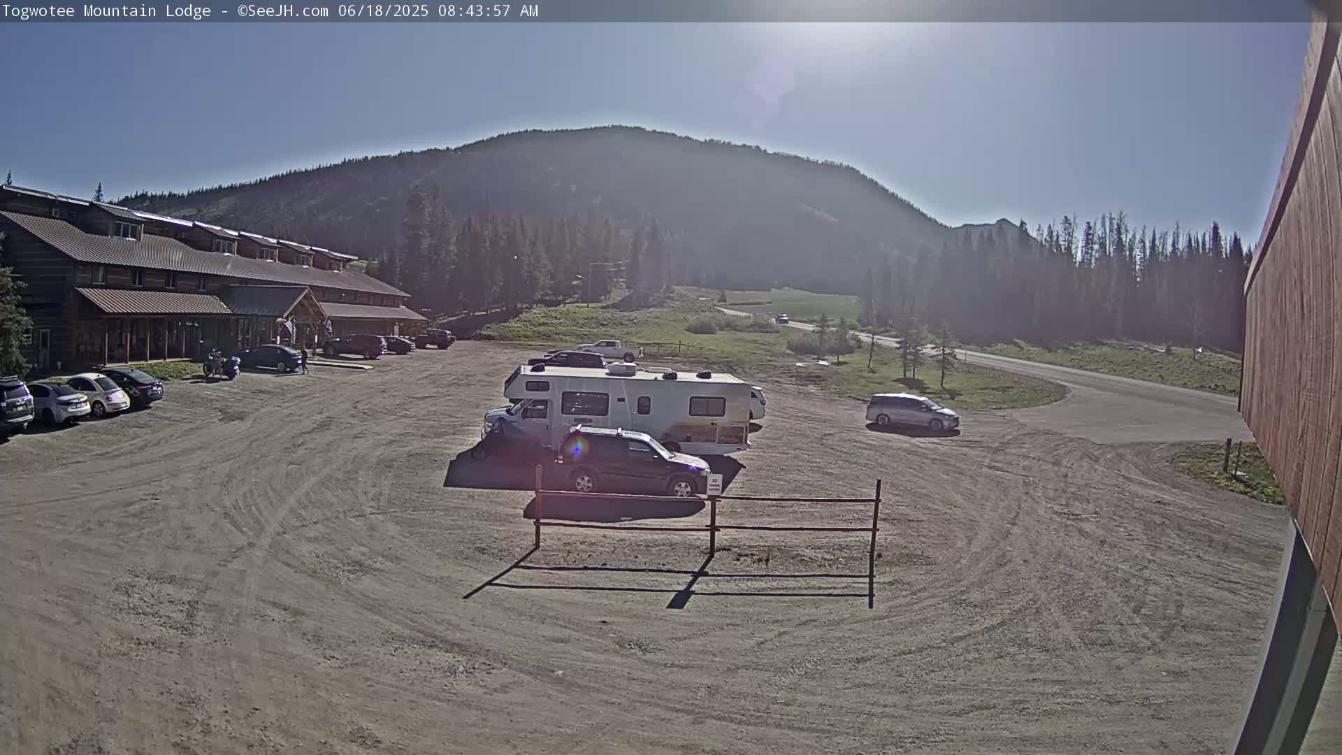A gravel parking lot with several cars and an RV is in front of a log building, all situated beneath a clear, sunny sky and a mountain range in the background.