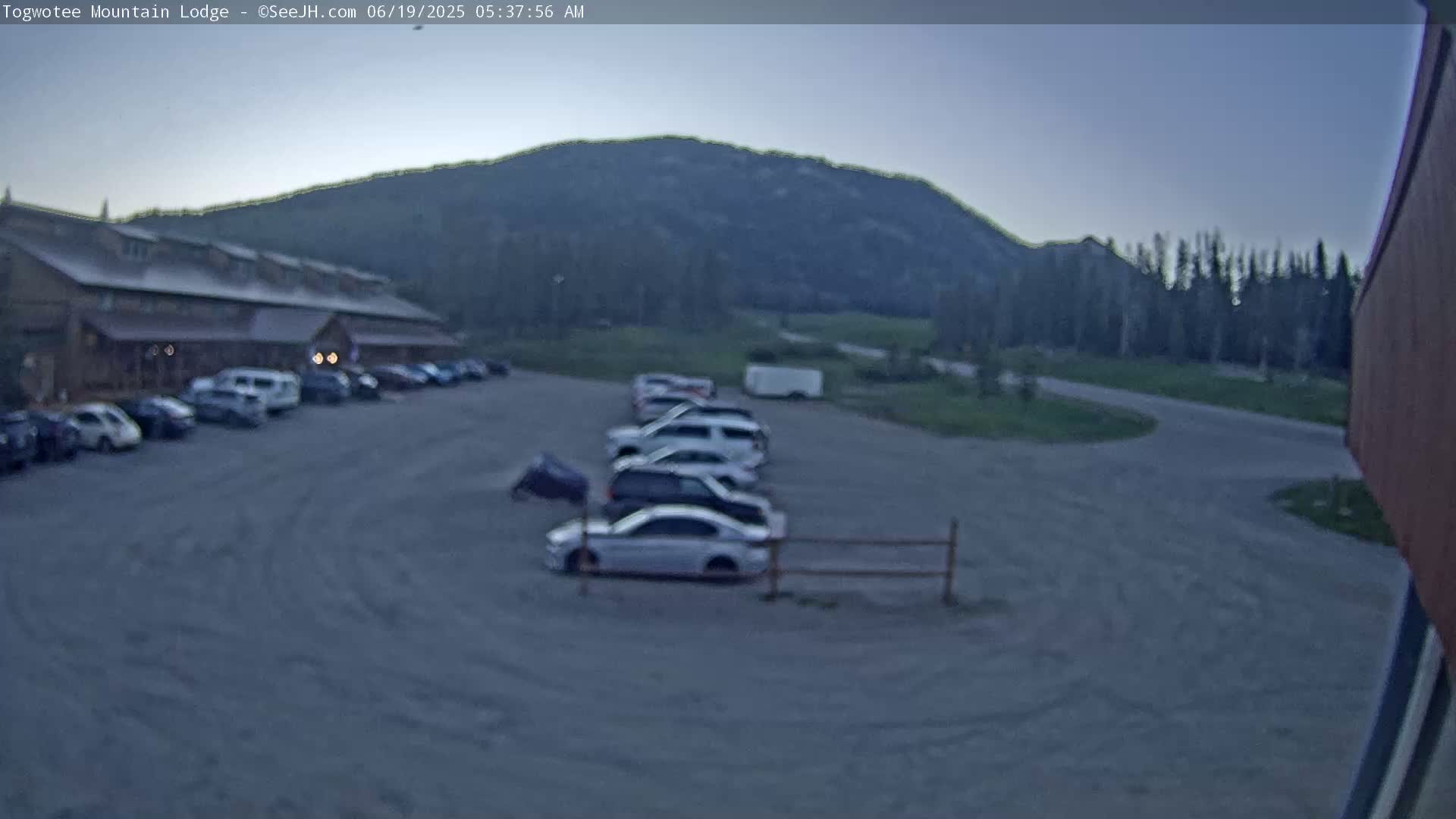 Several cars are parked in a gravel lot in front of a long, low building at the base of a mountain under a twilight sky.