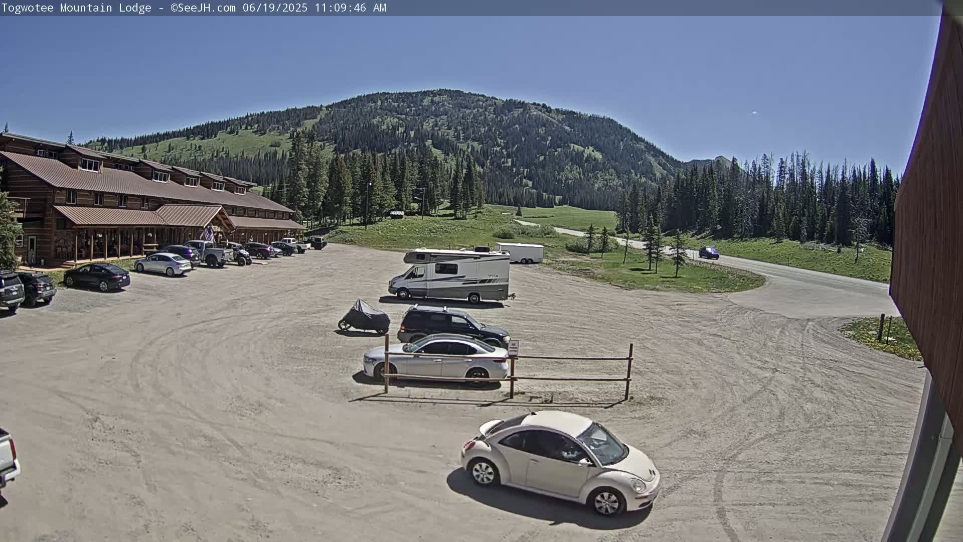 A parking lot with several cars and an RV in front of a log cabin lodge, situated against a backdrop of a mountain range under a clear, sunny sky.