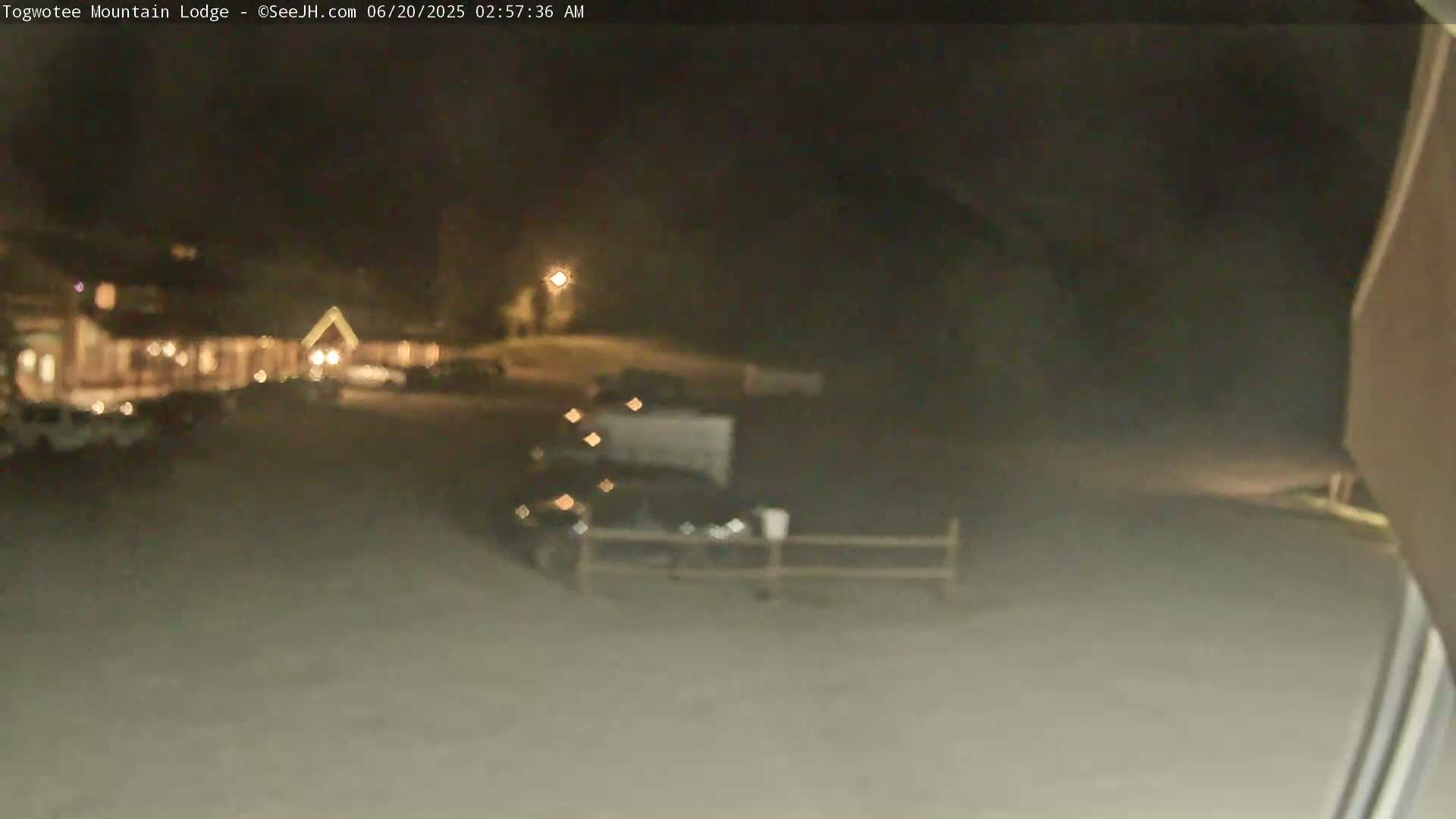 A nighttime, blurry view of a parking lot in front of a building with some cars parked, a low fence, and a streetlight.